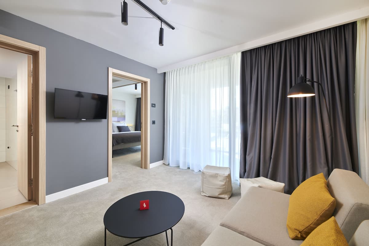 Una sala de estar moderna con un televisor de pantalla plana, un sofá con cojines color mostaza, una mesita redonda y un ventanal con cortinas.