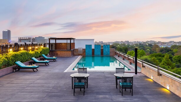 Un espace piscine aménagé sur le toit, avec des chaises longues et des tables, entouré de verdure et offrant une vue sur la ville.