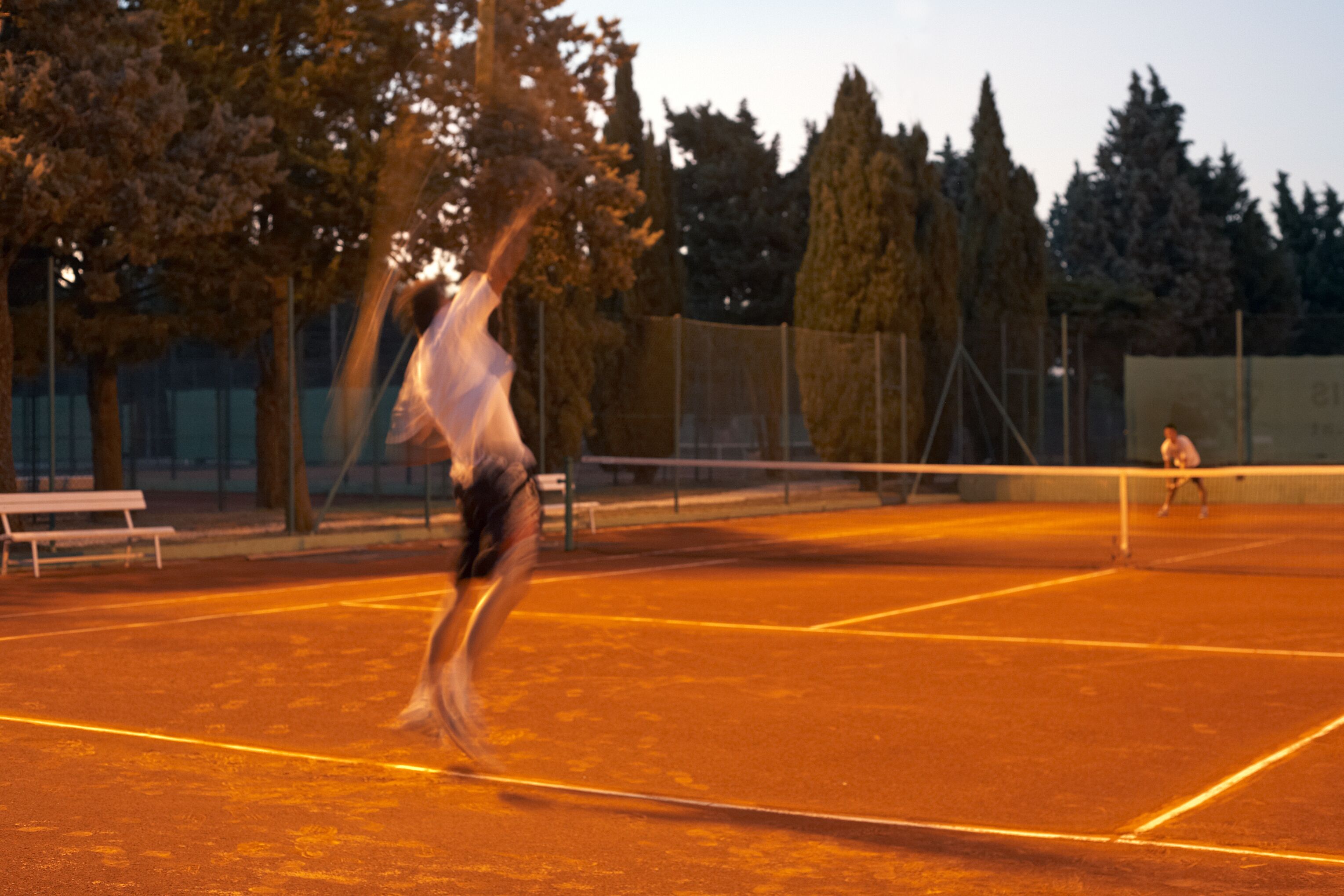 Una cancha de tenis con red, árboles y un banco.