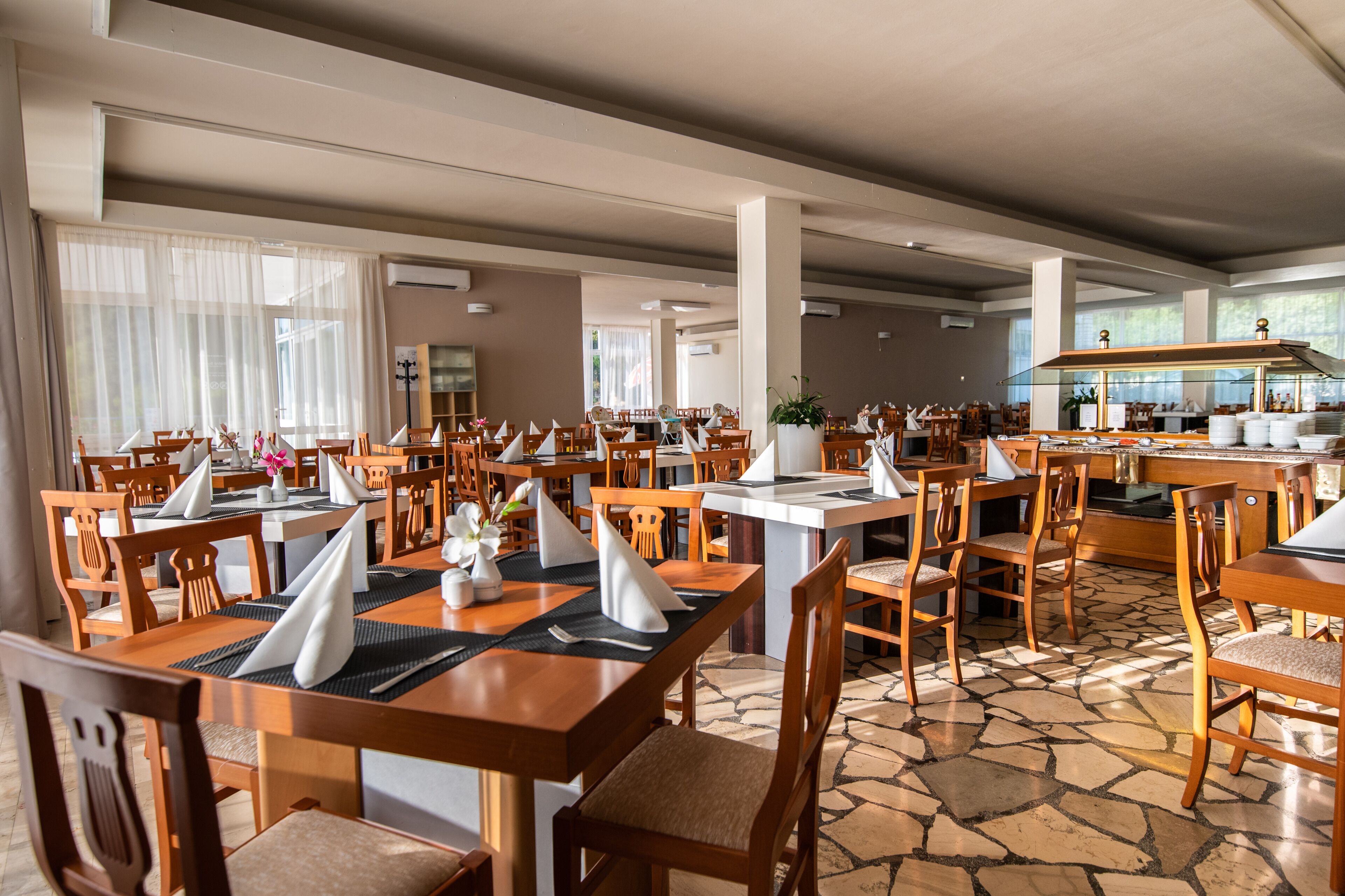Spaziosa sala da pranzo con diversi tavoli e sedie, ampie finestre e un'area cucina sullo sfondo.