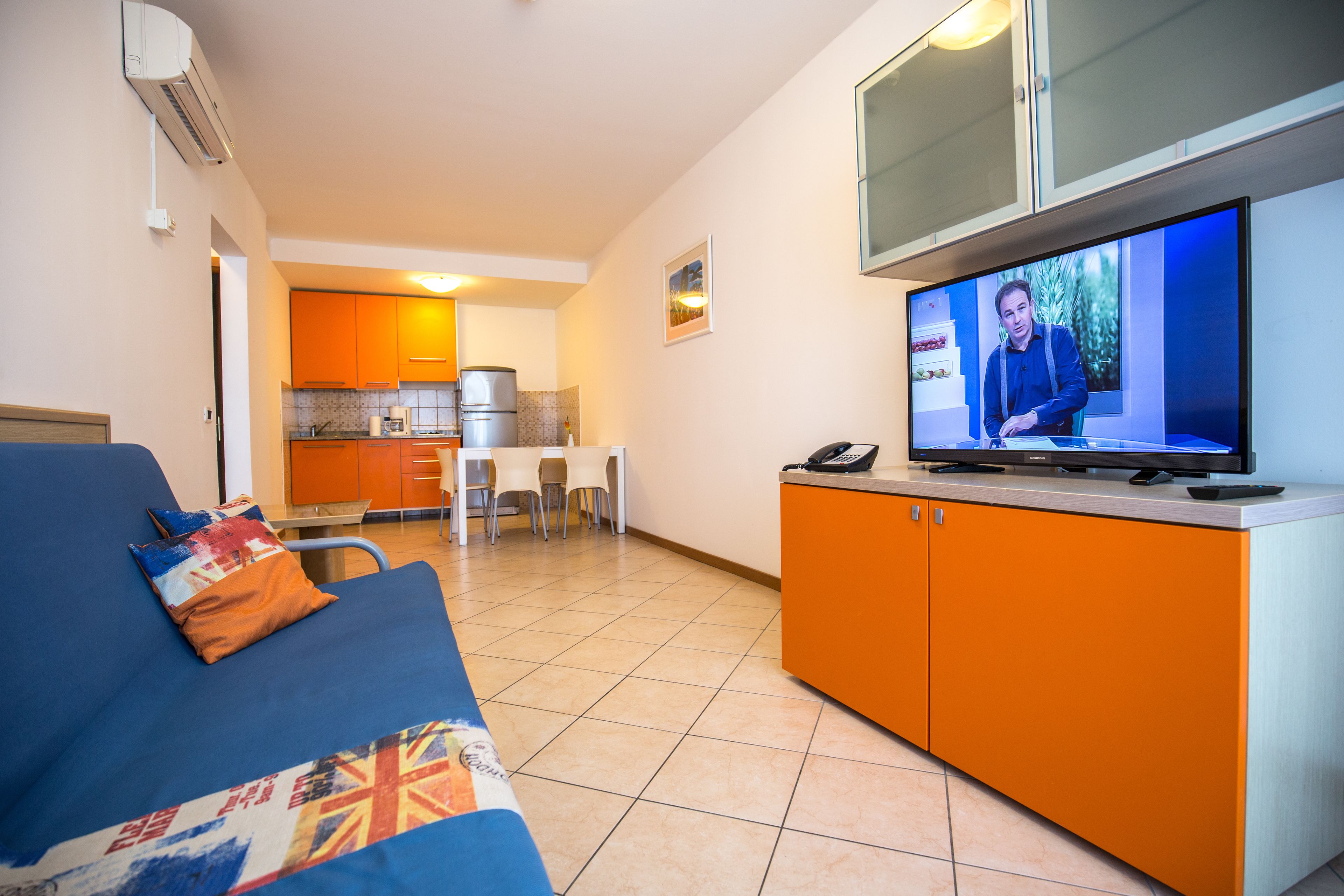 Uma sala de estar com um sofá azul, armários laranjas e uma televisão exibindo uma pessoa.