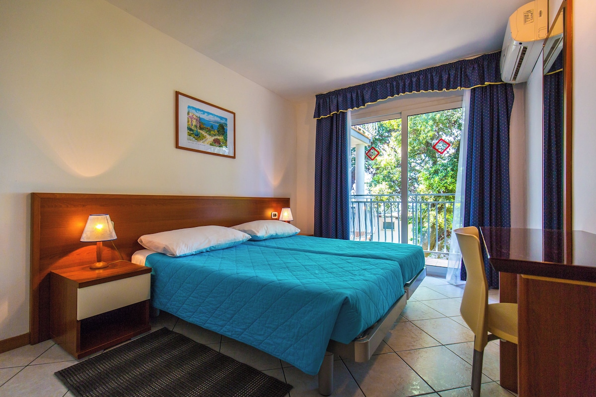 A hotel room with a bed, a desk, a chair, a nightstand, a lamp, a picture on the wall, and a view of the outdoors.
