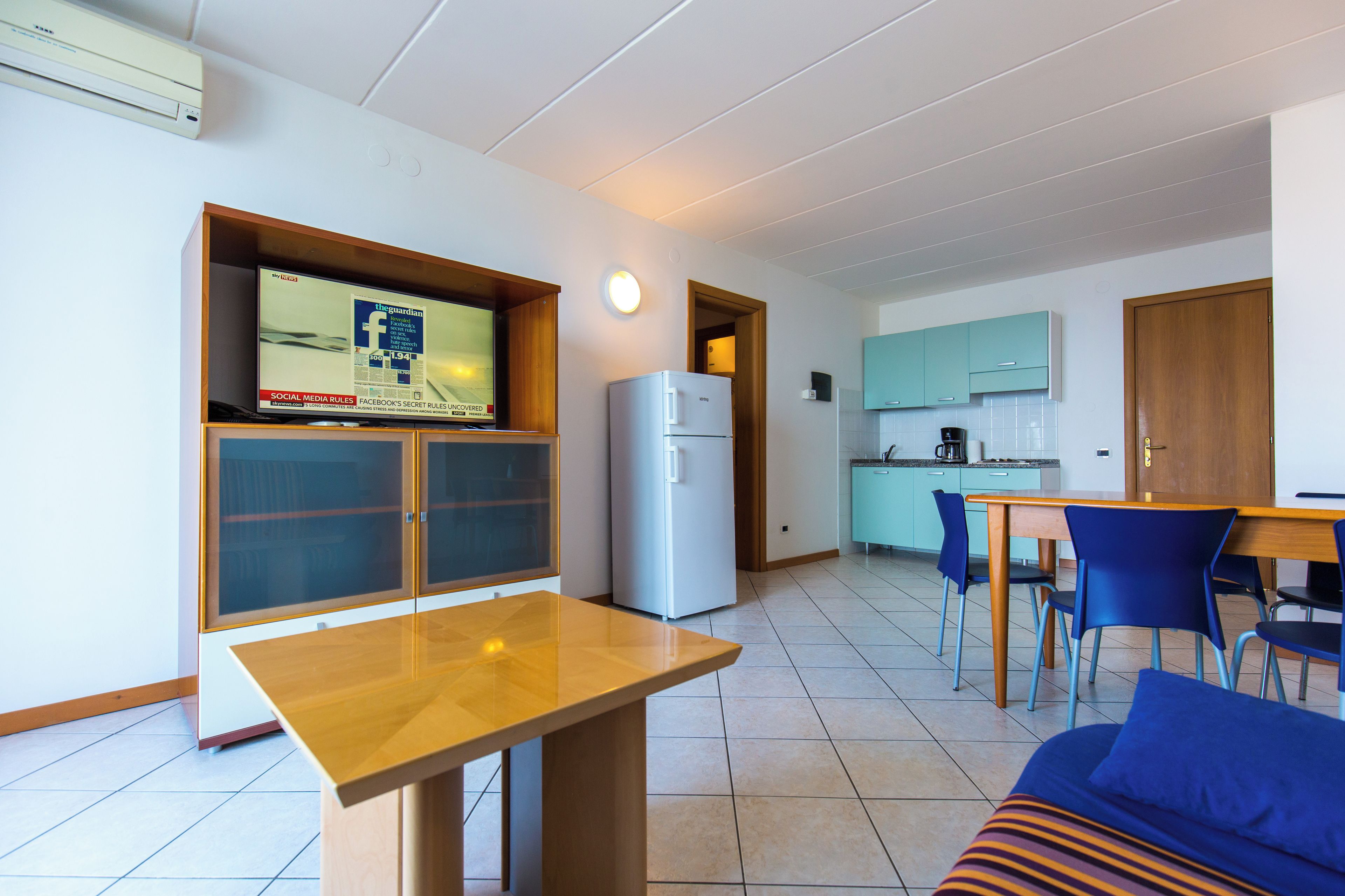 A compact kitchen with a refrigerator, a dining area with a table and chairs, and a television mounted on the wall.