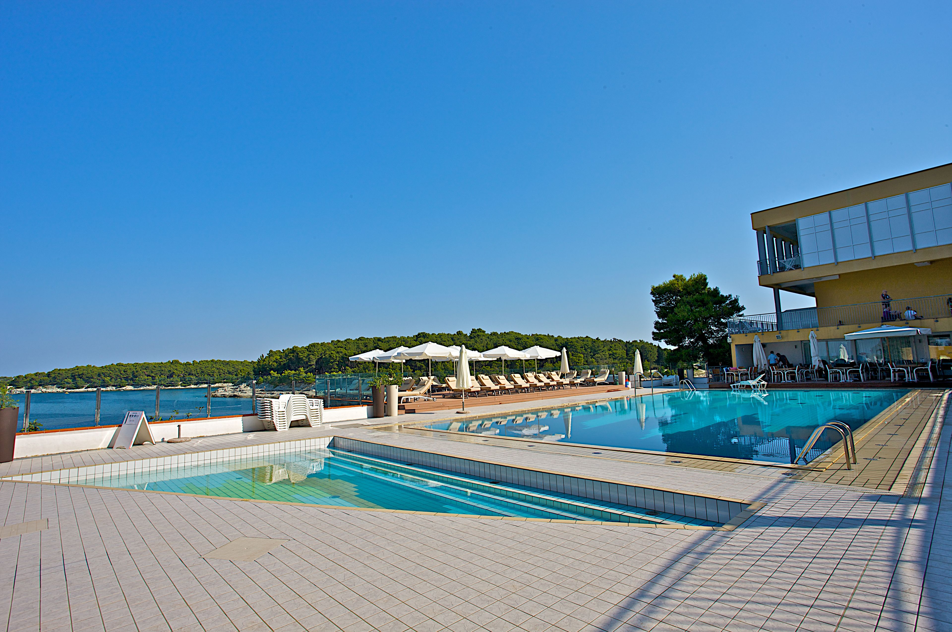 A swimming pool with sun loungers and umbrellas, a view of the sea, and a building with a balcony.