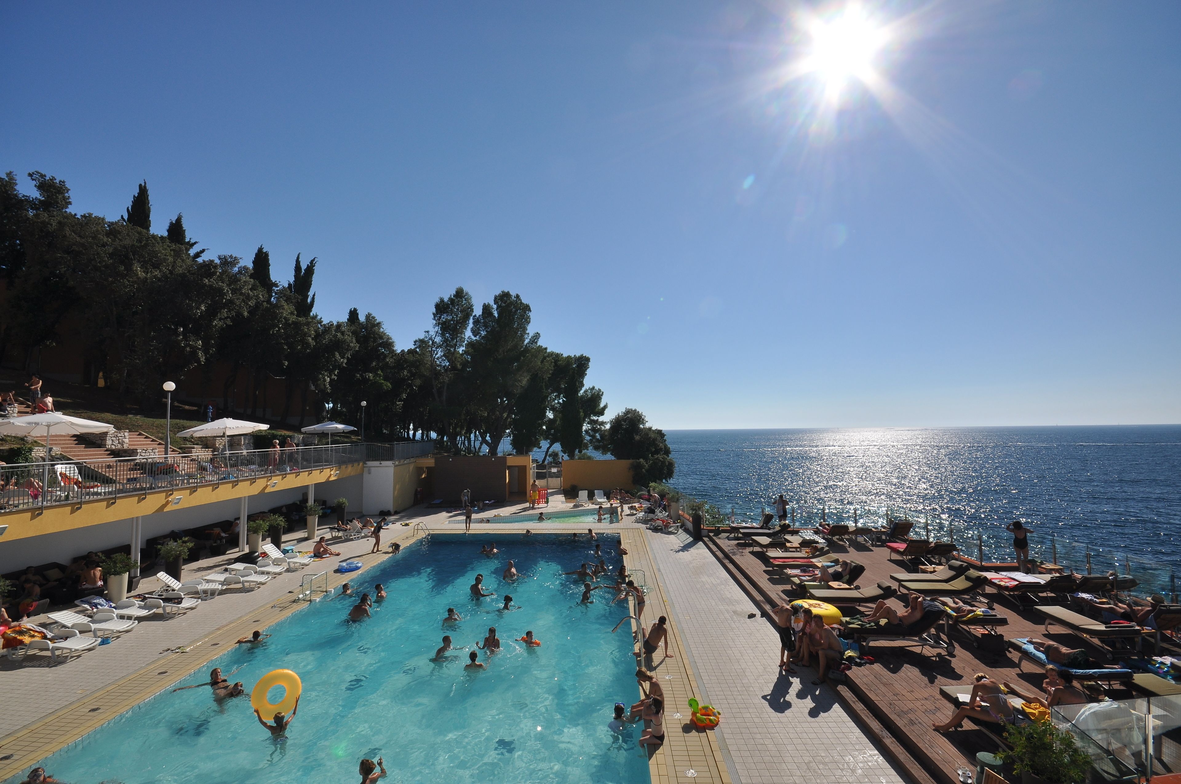 Ein Poolbereich mit Schwimmern und Liegestühlen, ein sonnendurchfluteter Himmel und Blick aufs Meer.
