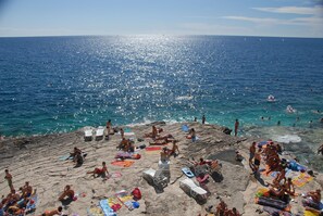 A beach with people sunbathing and swimming, rocky terrain, and clear blue water.