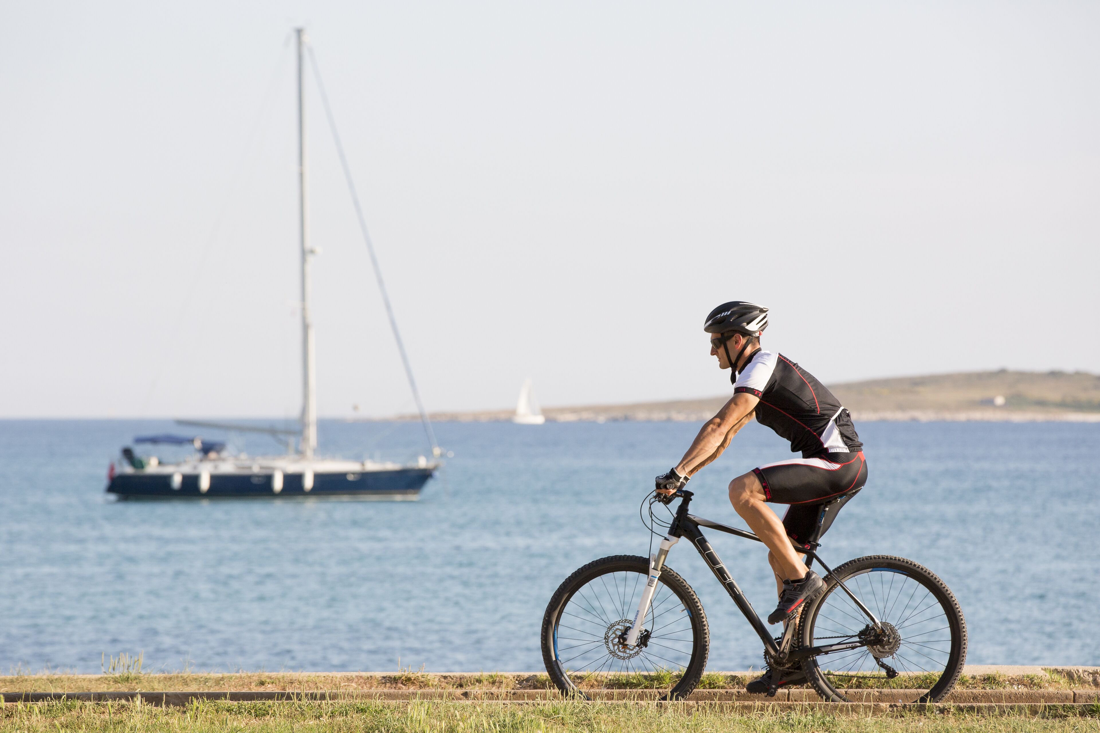 A cyclist riding a bike by the sea with a sailboat in the background.