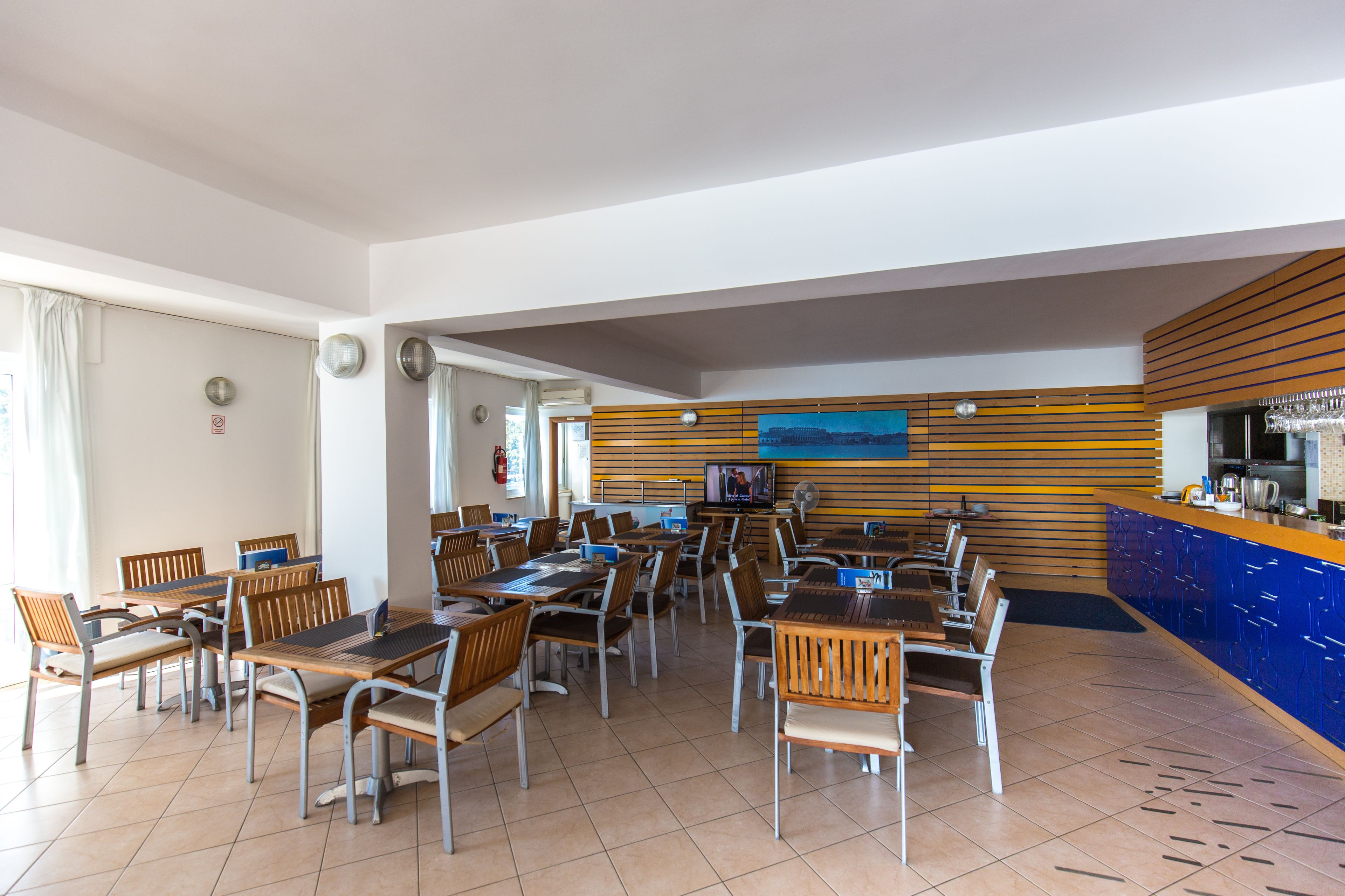A spacious dining area with multiple tables and chairs, a kitchen area, and a large window providing natural light.