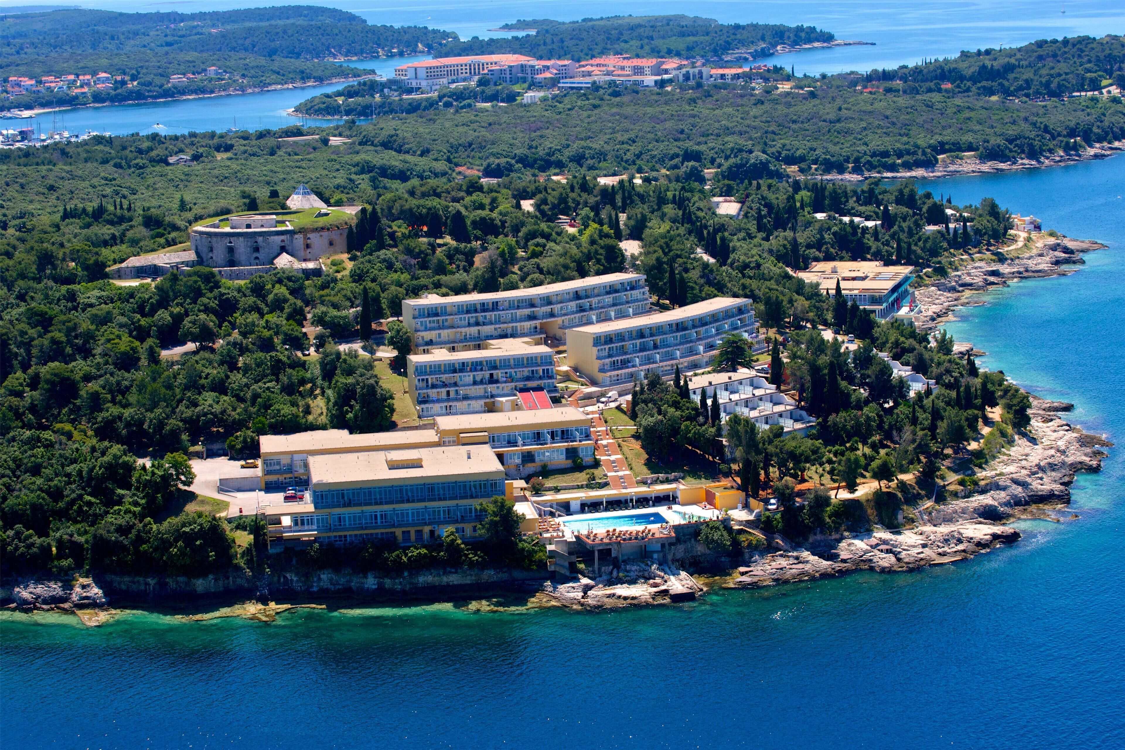An aerial view of a coastal hotel with multiple buildings, a swimming pool, and surrounding greenery.
