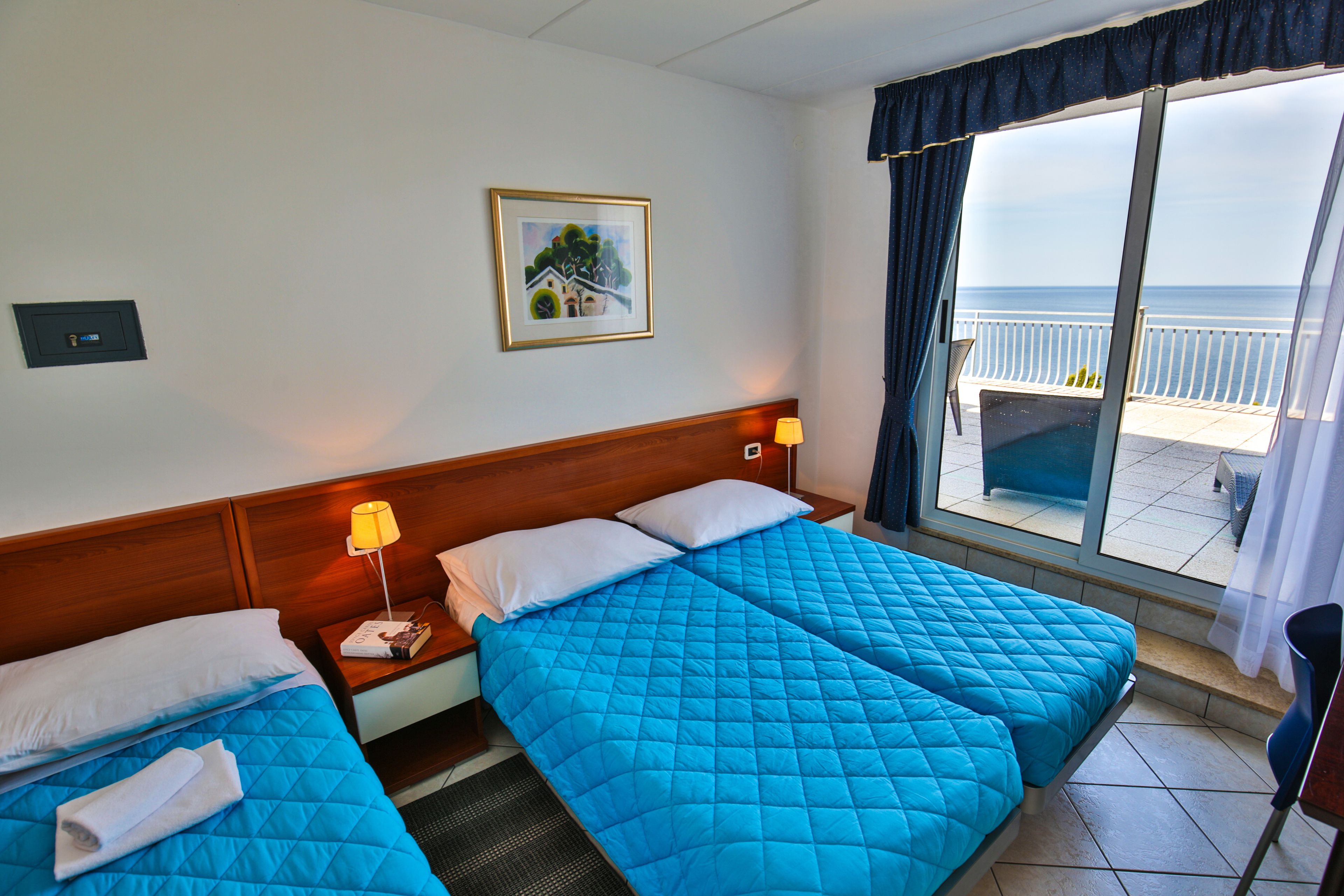 Une chambre d’hôtel avec deux lits, une tête de lit en bois, une table de chevet avec une lampe, un tableau encadré au mur et une vue sur la mer depuis un balcon.