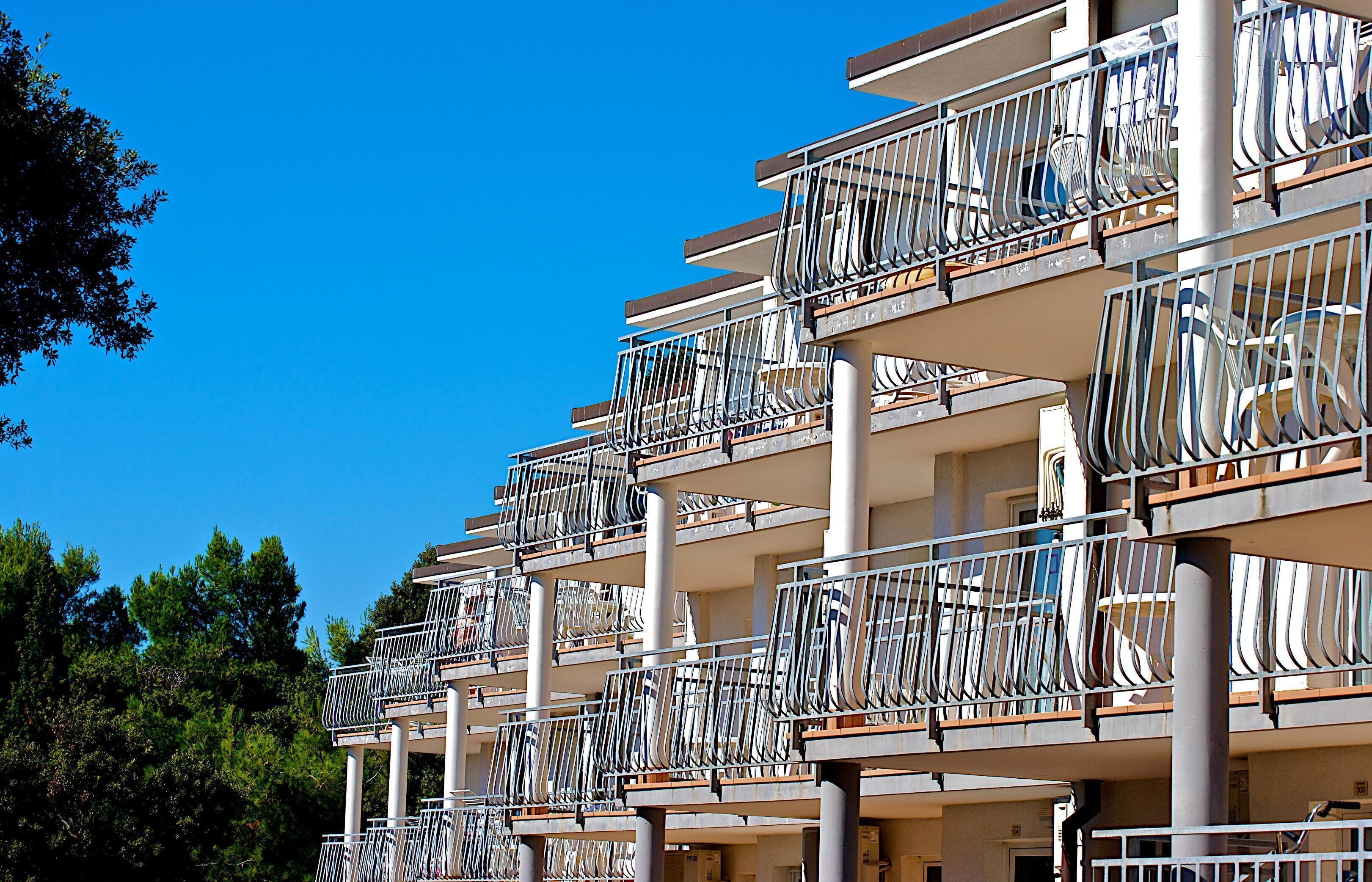 Een hotel met meerdere verdiepingen, voorzien van balkons met metalen hekwerk en een helderblauwe lucht op de achtergrond.