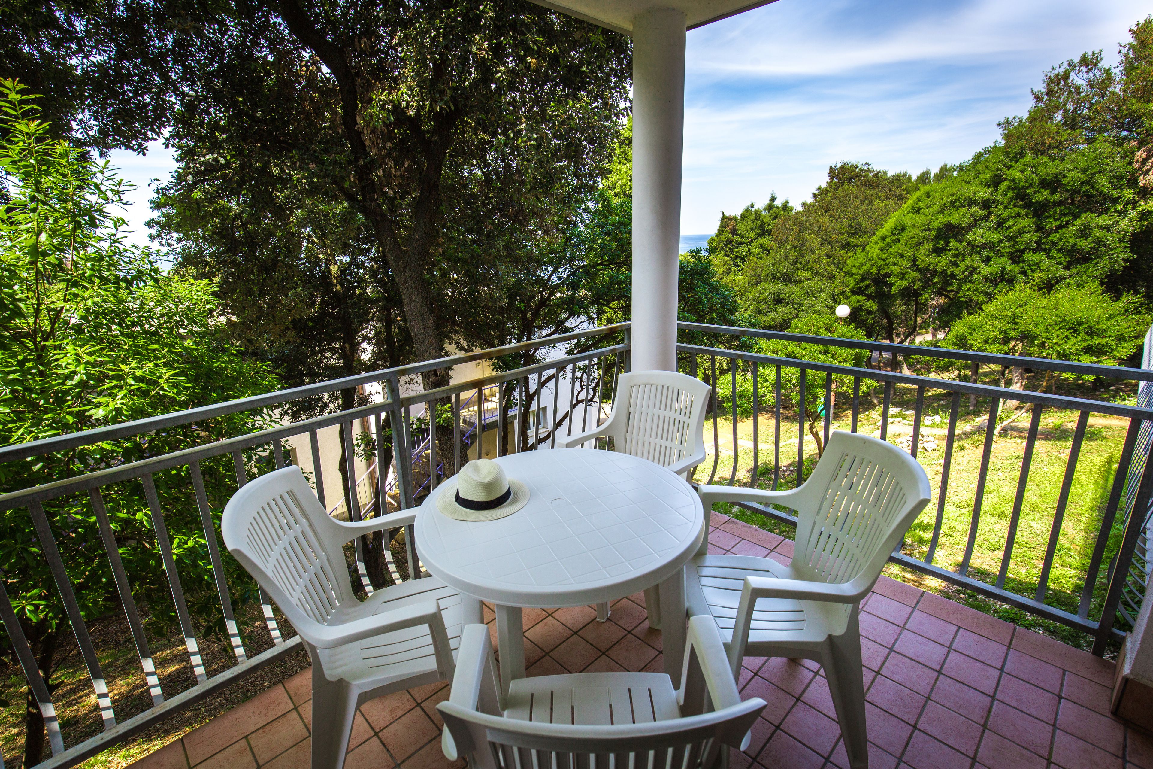 Un balcon avec une table ronde et quatre chaises blanches, entouré d’une rambarde en métal.