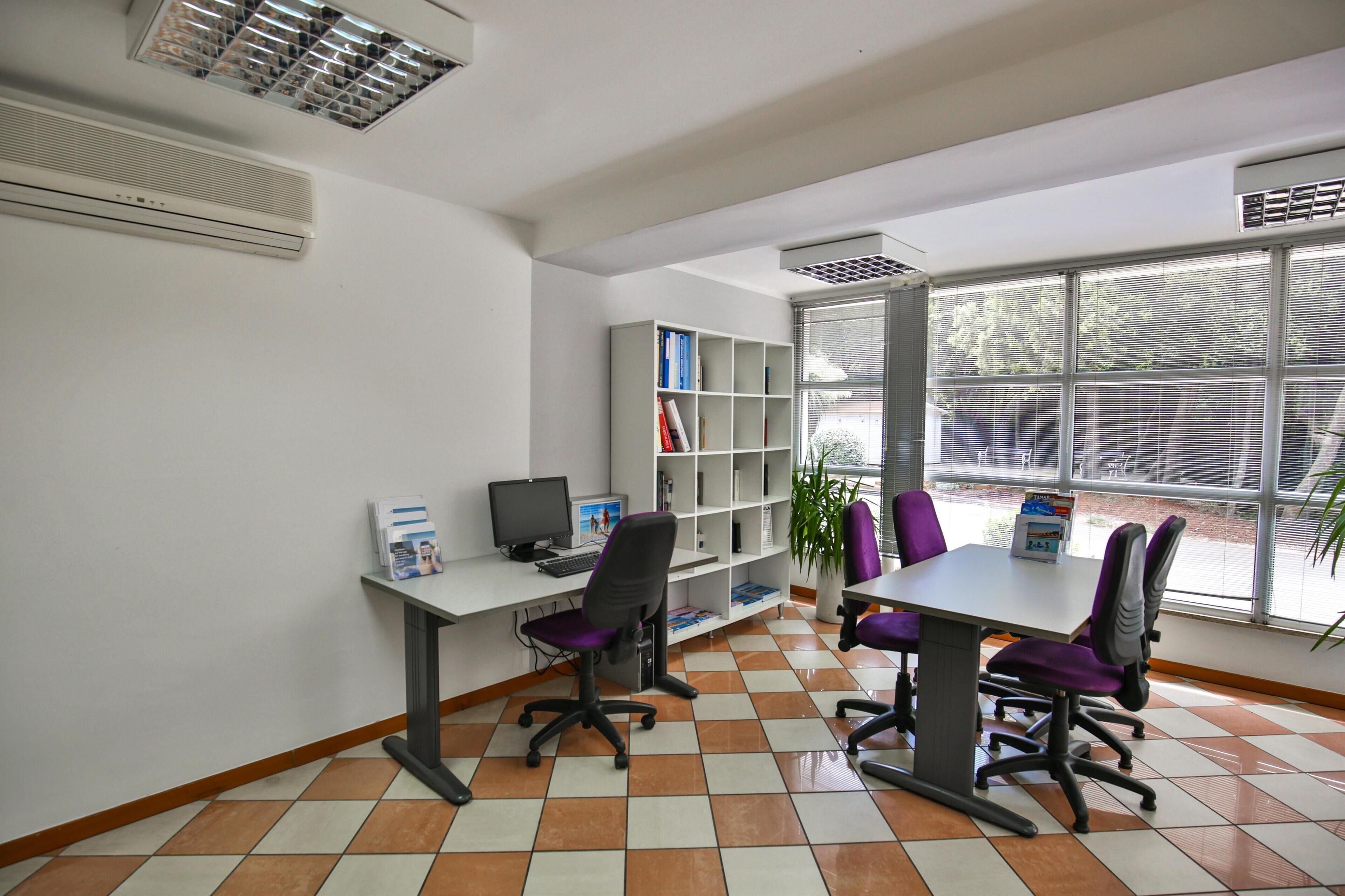 Une salle de conférence avec une table blanche, des chaises violettes, un ordinateur et une bibliothèque.