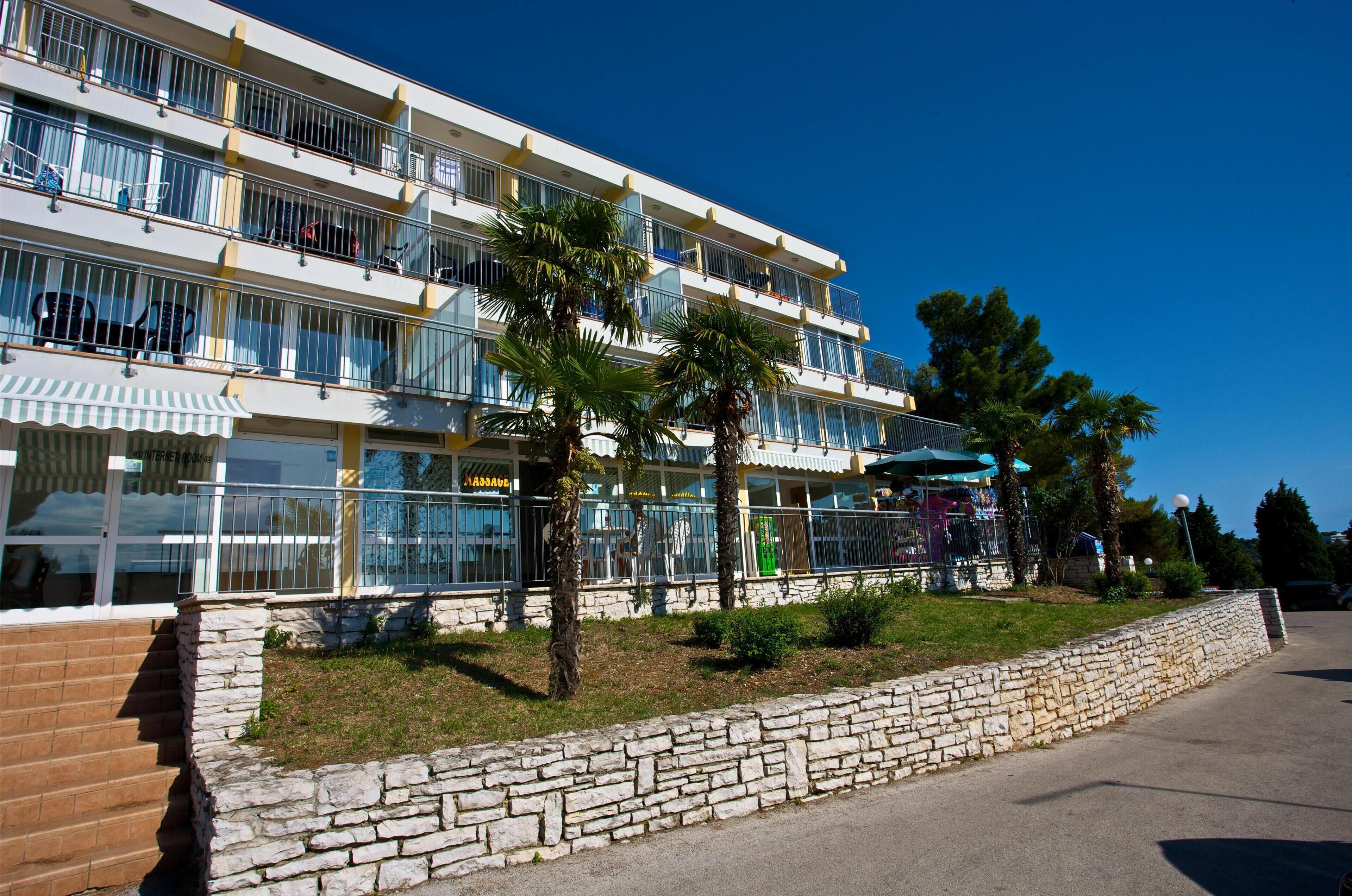 A multi-story hotel with balconies, a stone retaining wall, and palm trees.