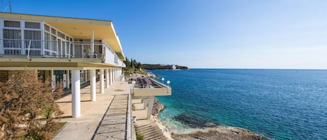 Un edificio costero con balcón que da al mar, una escalera hacia la playa y vistas despejadas de la costa.