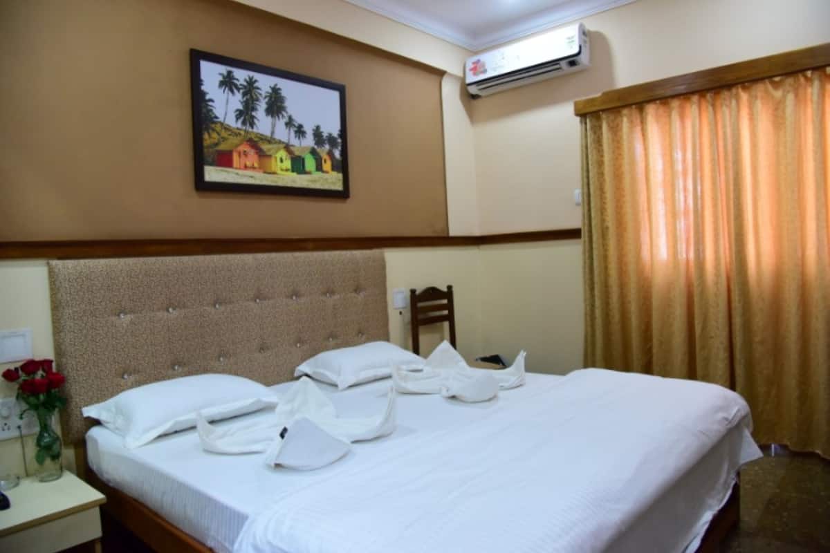 A hotel room with a bed, a nightstand with a vase of flowers, a painting on the wall, and an air conditioning unit.
