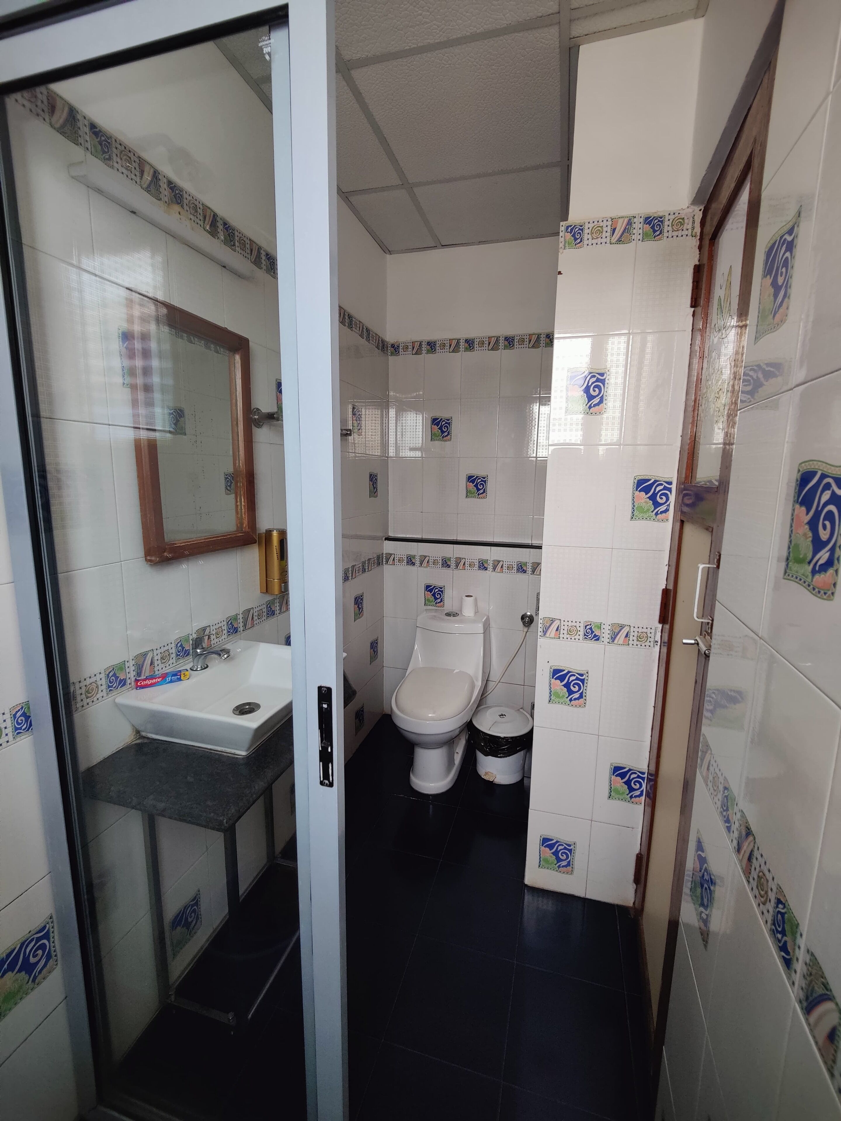 A bathroom with a white sink, a toilet, and a trash bin. The walls are tiled with a decorative pattern.