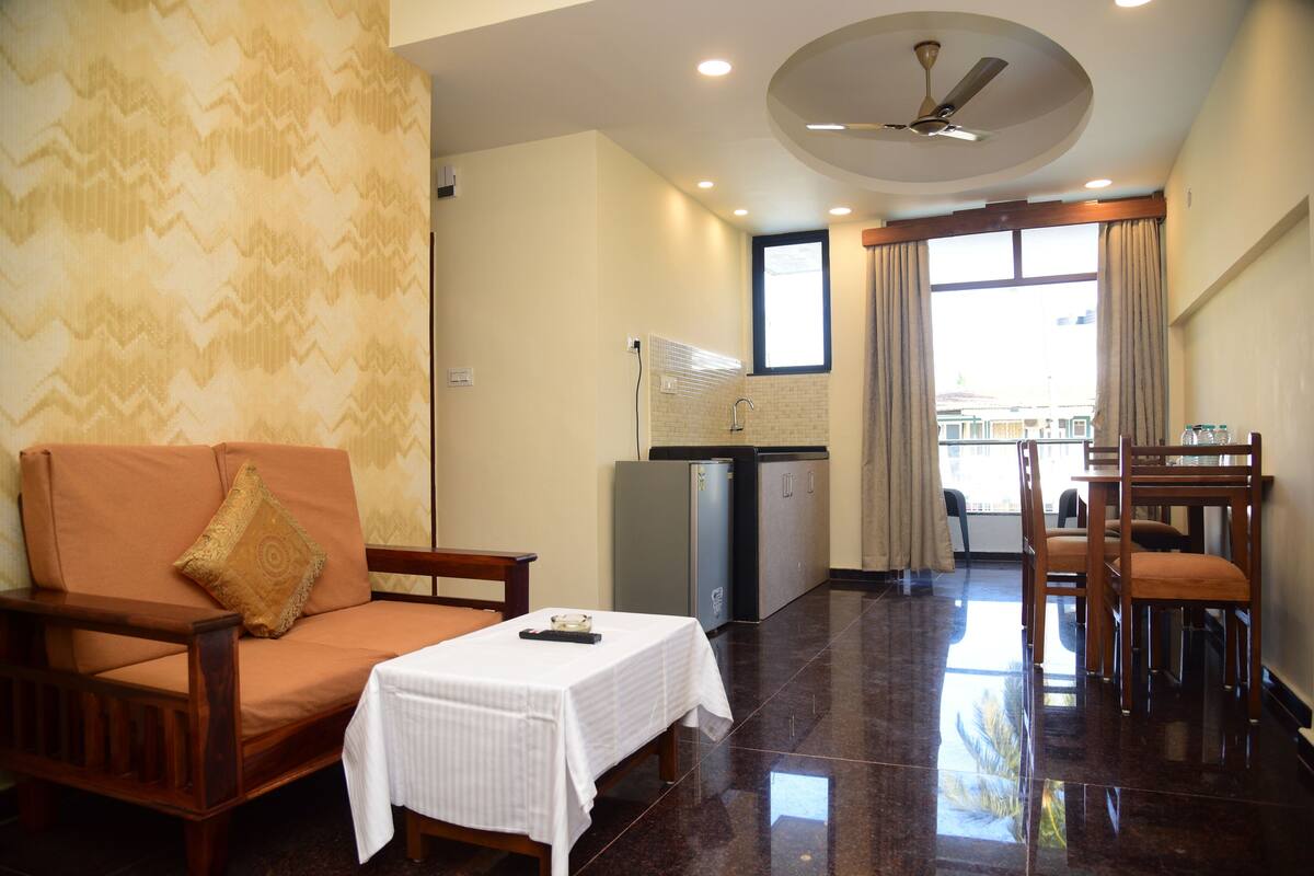 A hotel room with a sofa, a small table, a dining table with chairs, a ceiling fan, a window with curtains, and a minibar.