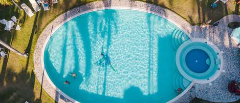 An aerial view of a circular swimming pool with a stone edge, surrounded by lounge chairs and umbrellas, and palm trees.