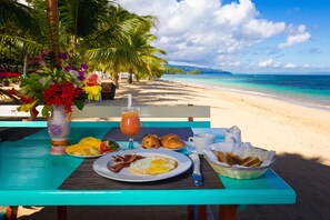 Une table de petit-déjeuner avec des œufs, du bacon, des toasts, des croissants et du jus, installée sur une plage bordée de palmiers et offrant une vue sur l’océan.