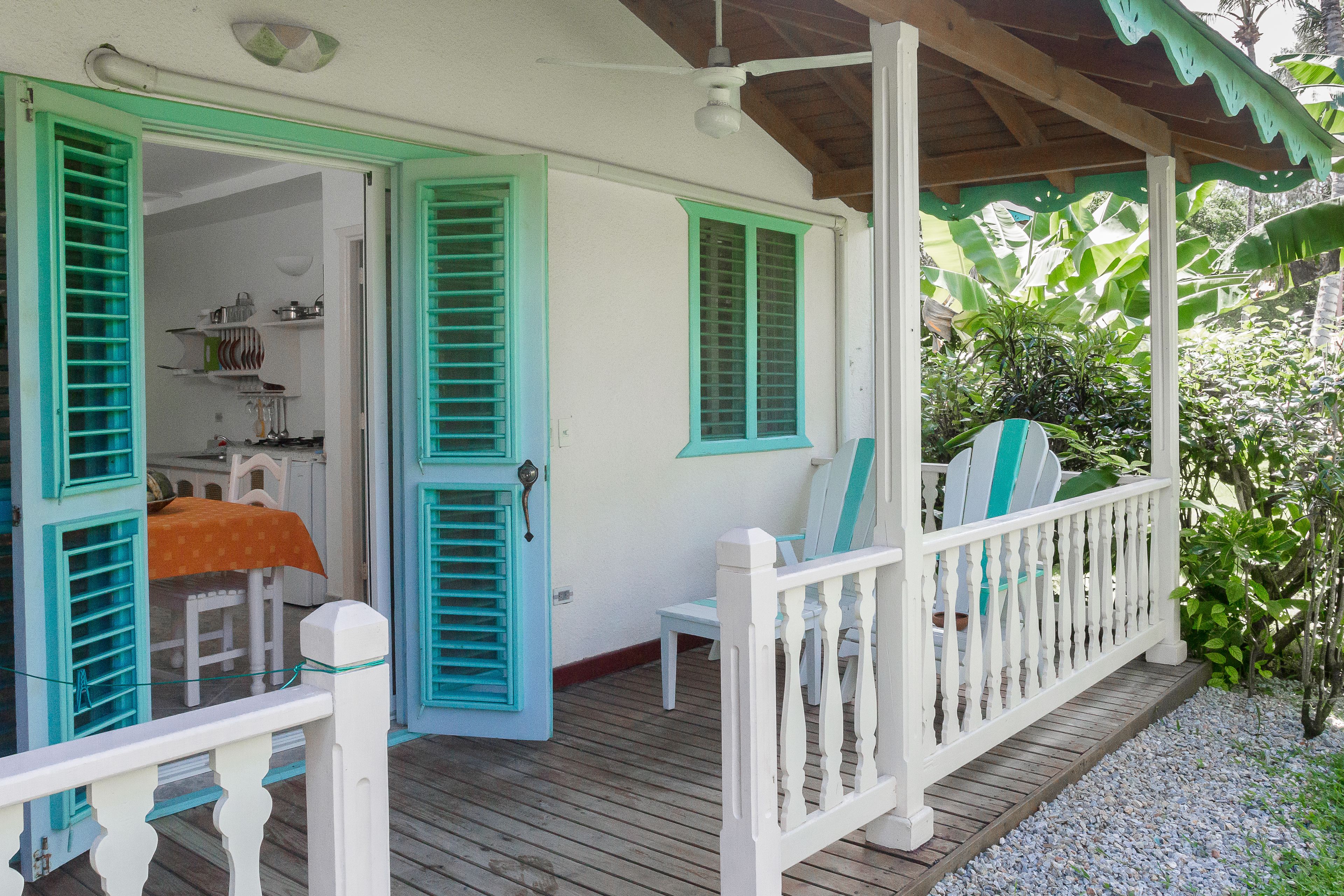 Una terraza de madera con barandal blanco, puertas abiertas con contraventanas verdes y una zona de estar cubierta con una tabla de surf.