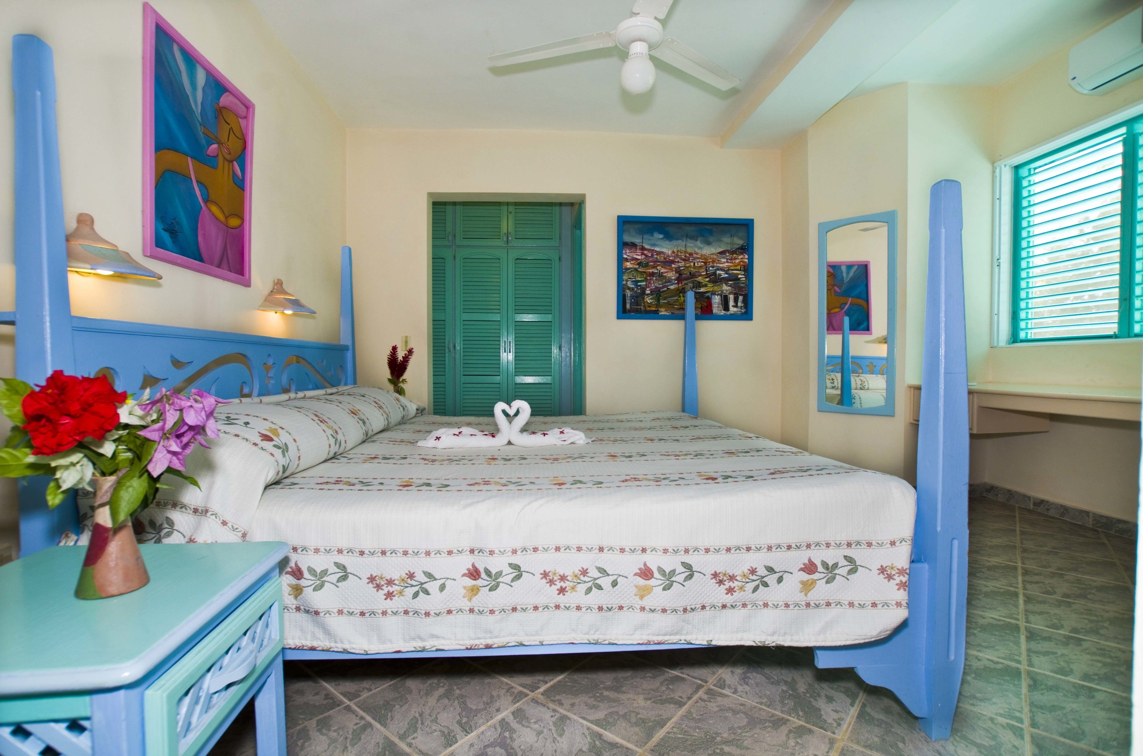 A bedroom with a bed, a nightstand, a vase with flowers, a ceiling fan, and a window with blinds.