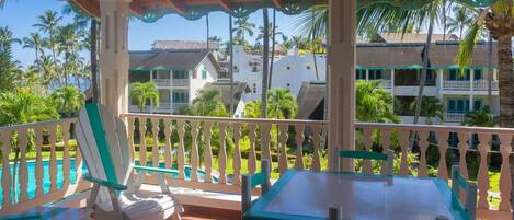 A covered outdoor area with a pool, white and green furniture, and a view of a building with balconies and palm trees.