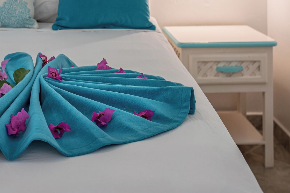 Ein ordentlich bezogenes Bett mit einer türkisblauen Decke und rosa Blumen, ein weißes Kopfteil und ein Nachttisch mit einer Lampe.