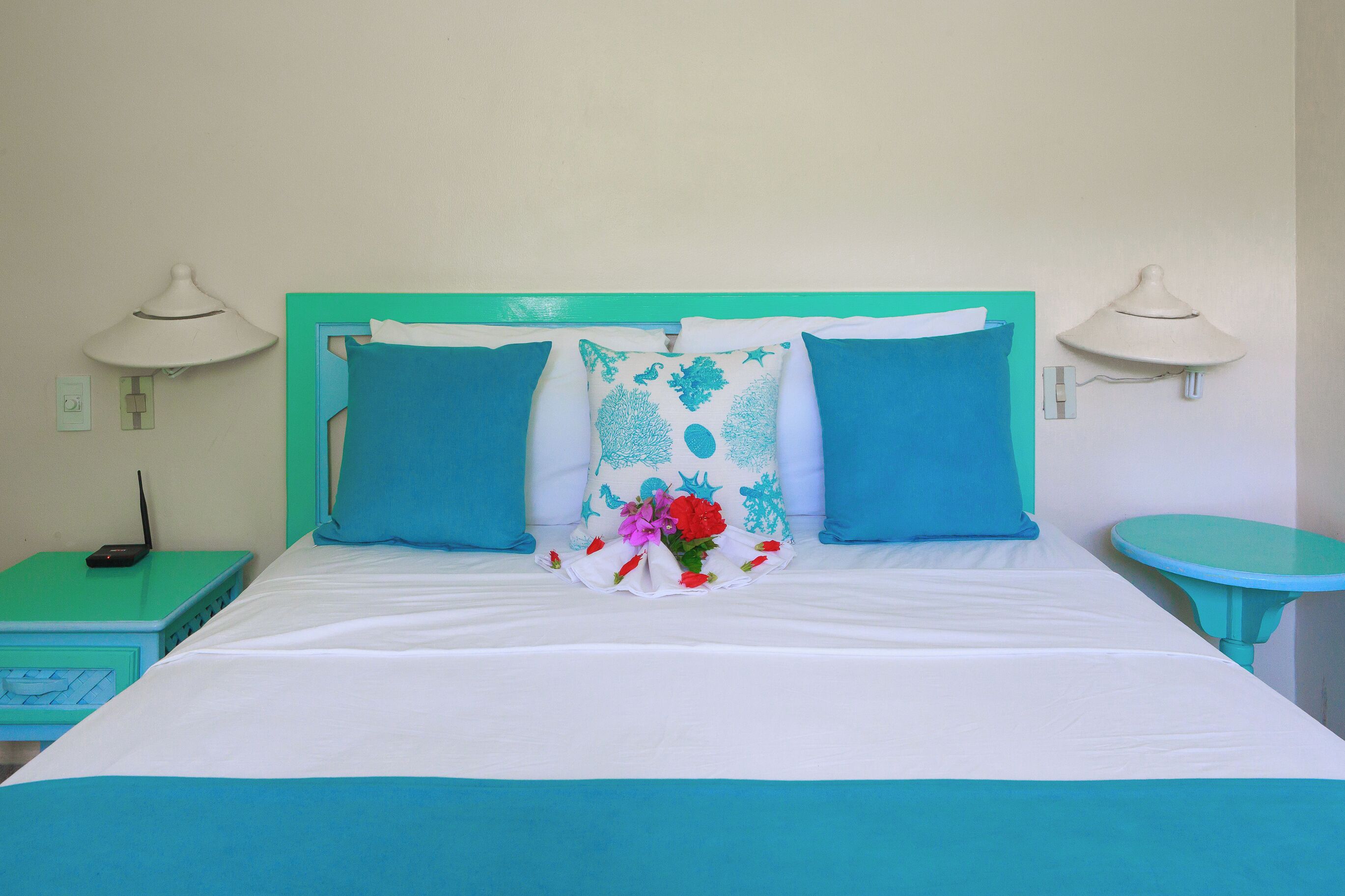 A neatly made bed with a white and teal color scheme, two matching pillows, a decorative pillow with a floral design, and a small teal side table with a lamp.