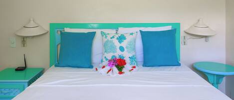 A neatly made bed with a white and teal color scheme, two matching pillows, a decorative pillow with a floral design, and a small teal side table with a lamp.