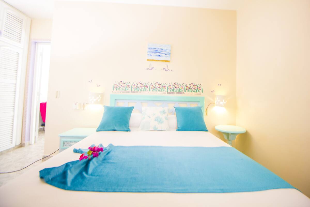 Ein Schlafzimmer mit einem Bett, zwei Nachttischen, einem Wandbild und einem Fenster mit Jalousien.