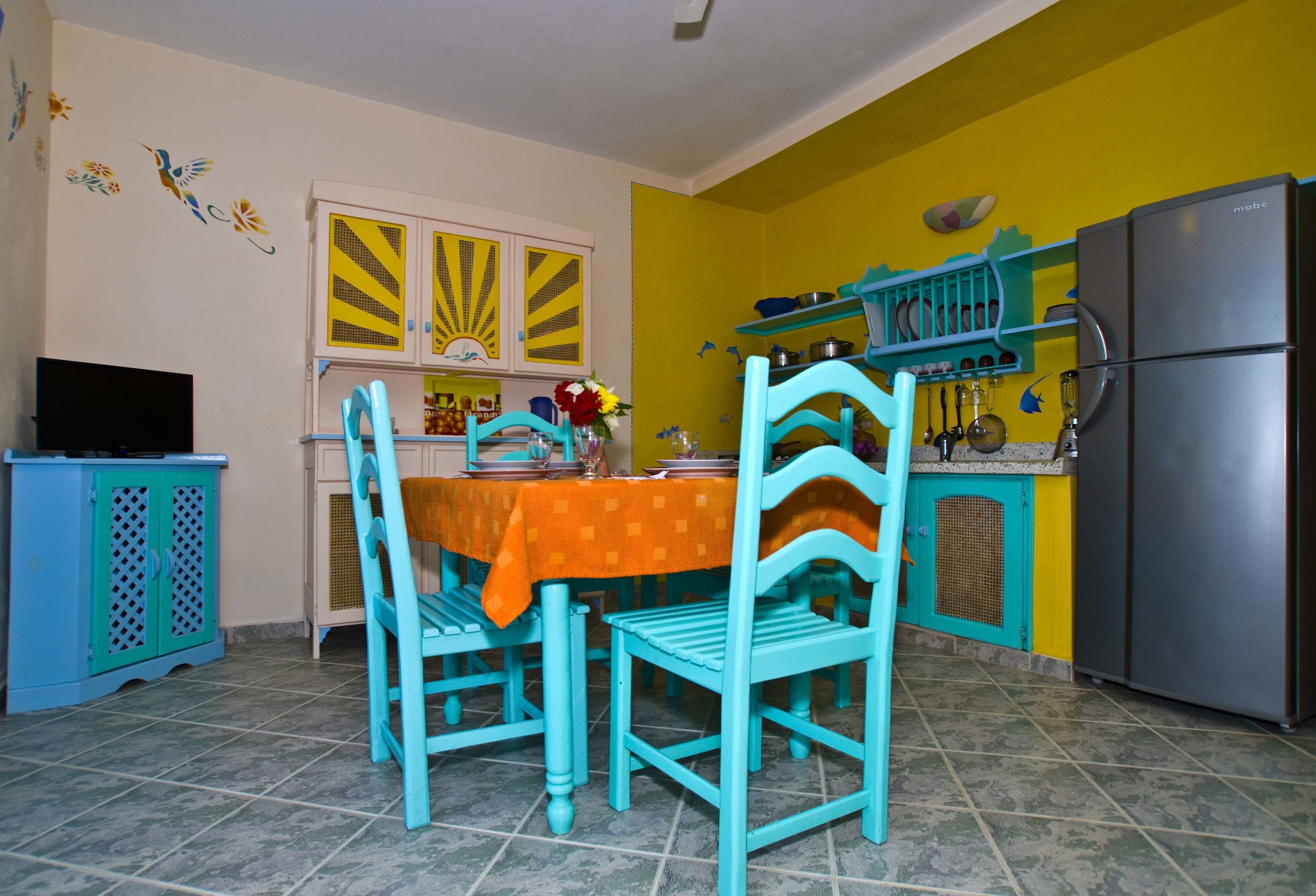 Una cocina con sillas azul verdosas, una mesa con mantel naranja y un refrigerador.