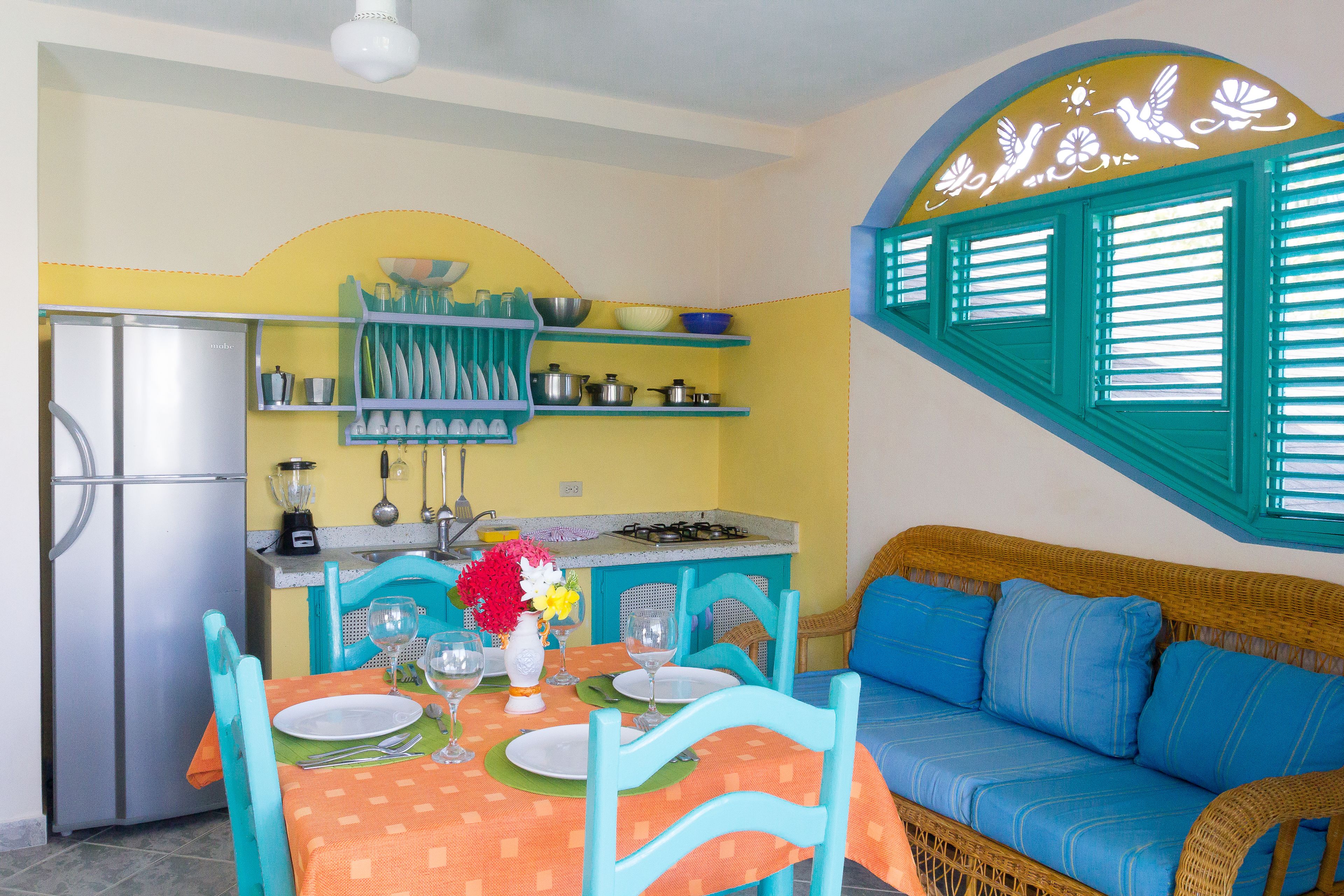 Cocina compacta con refrigerador, estufa y repisas con vajilla. Hay una mesa de comedor con sillas y un sofá con cojines.
