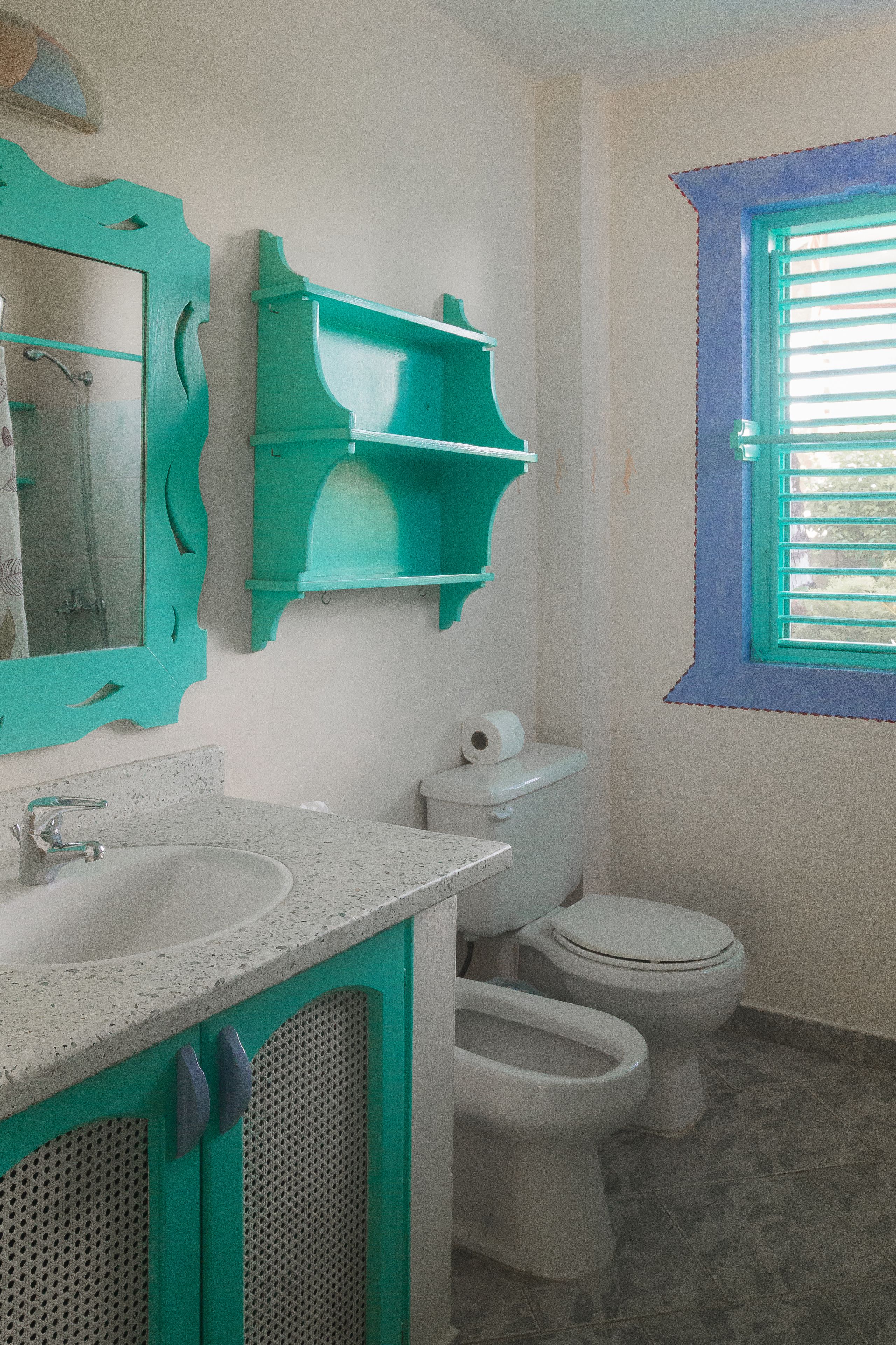 Un baño con un lavamanos blanco, dos inodoros, un espejo con marco verde azulado y una ventana con contraventanas azules.