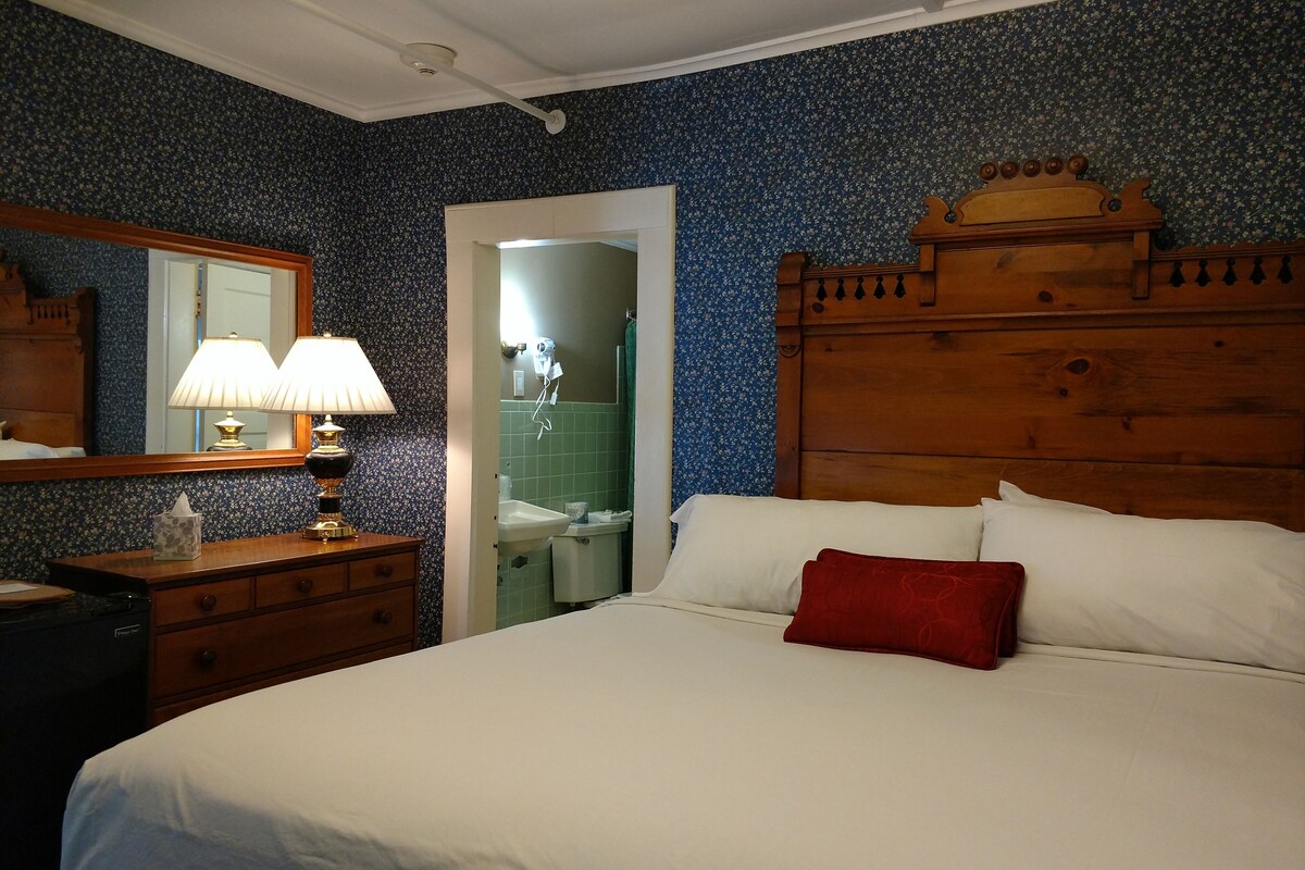 Un dormitorio con cabecera de madera, cama con ropa de cama blanca y almohada roja, mesita de noche con lámpara, cómoda y espejo.