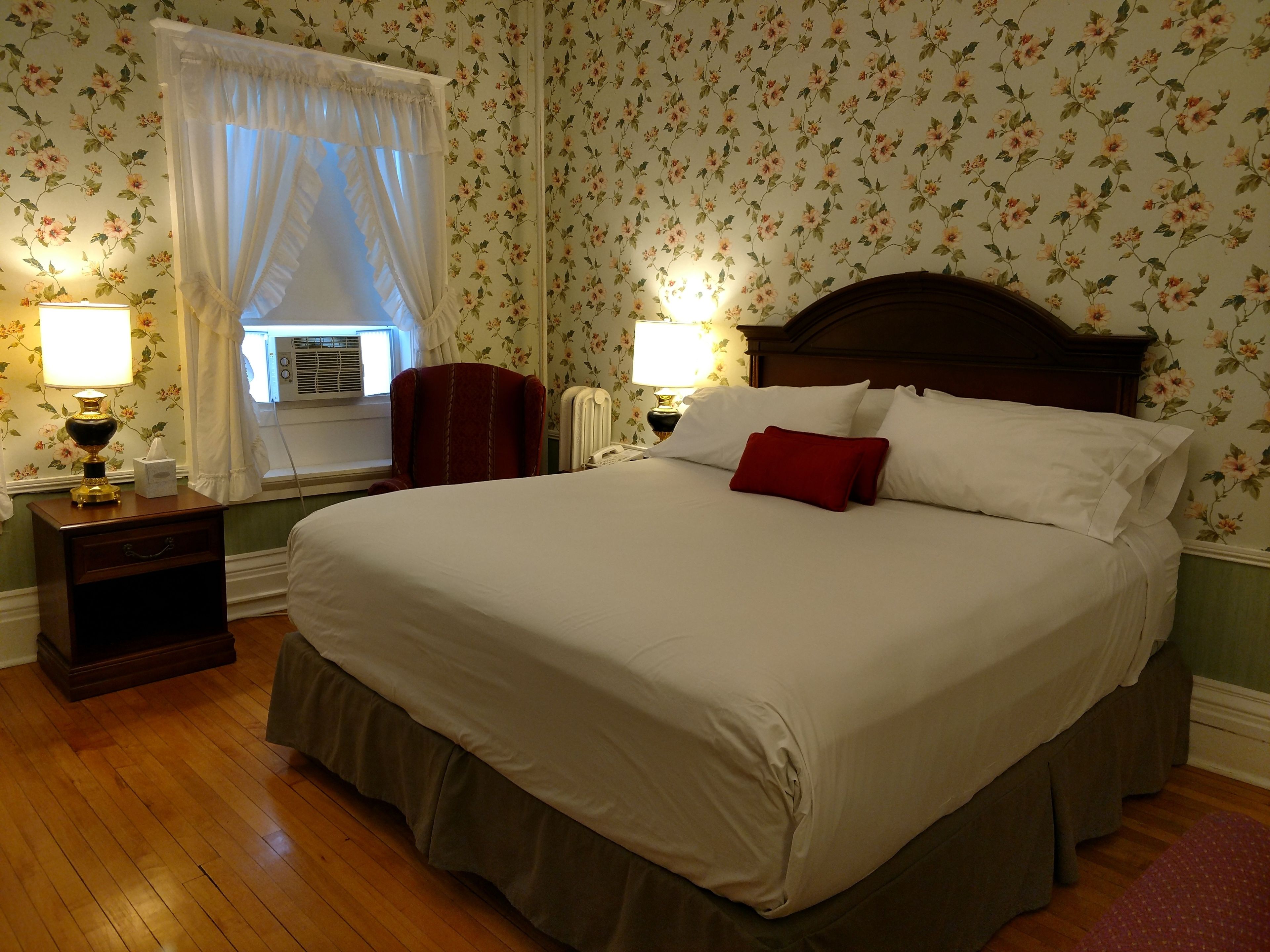 Una cama bien hecha con sábanas blancas y un cojín rojo, flanqueada por dos mesitas de noche con lámparas, en una habitación con papel tapiz floral y suelo de madera.