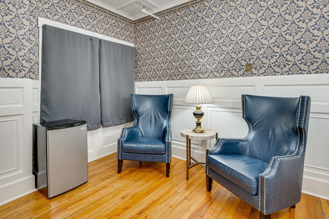 A room with two blue leather chairs, a small table with a lamp, a refrigerator, and patterned wallpaper.