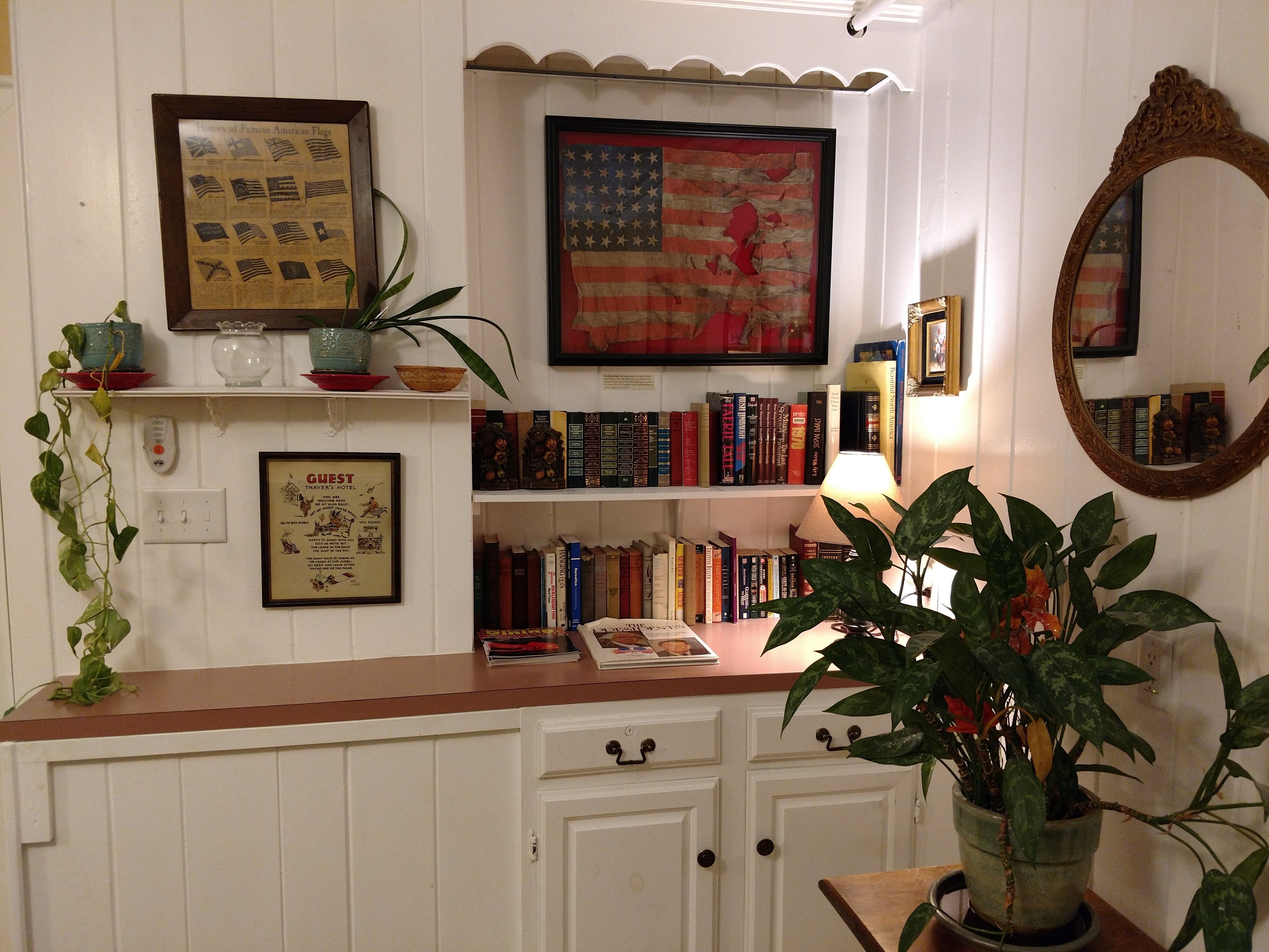 Quarto com parede branca, uma bandeira americana emoldurada, um espelho e uma estante de livros.