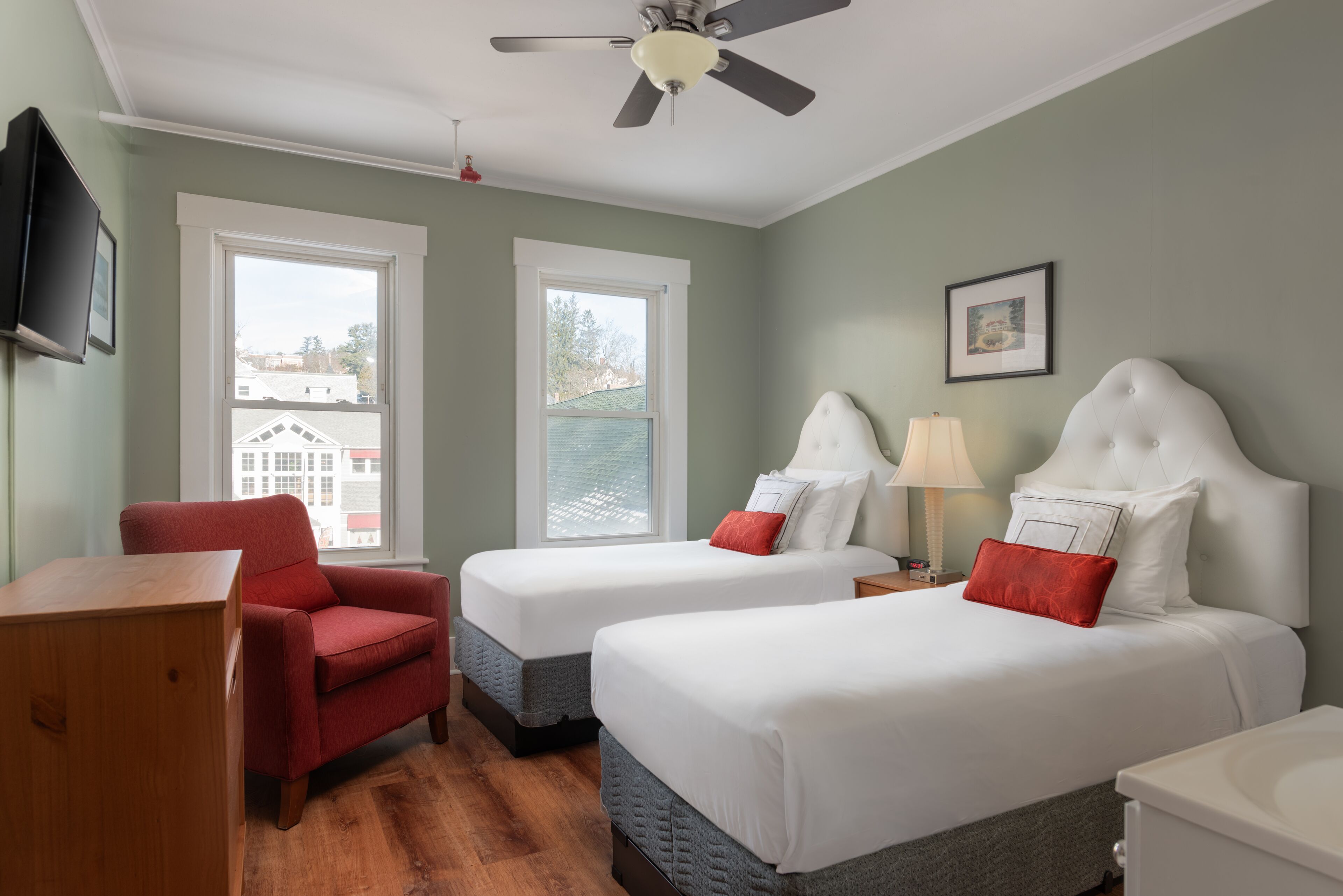 Quarto de hotel com duas camas, uma poltrona vermelha, uma escrivaninha de madeira e um ventilador de teto.