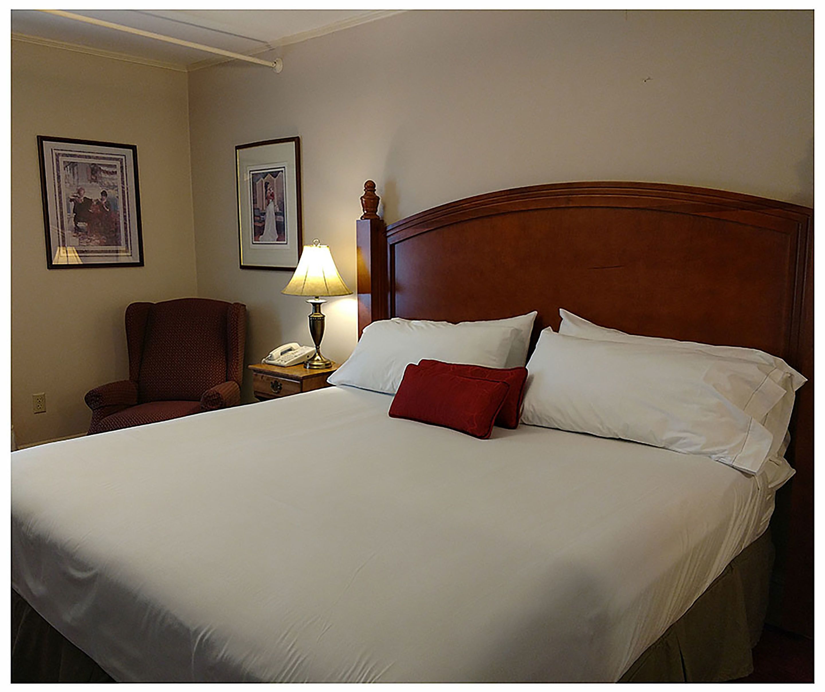 A neatly made bed with white linens and a red pillow, a wooden headboard, a bedside lamp, and two framed pictures on the wall.