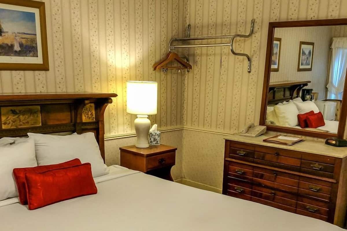 A hotel room with a bed, a nightstand with a lamp, a dresser, a mirror, and a hanging coat rack.
