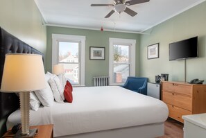 A hotel room with a bed, a desk, a chair, a dresser, a TV, and two windows.
