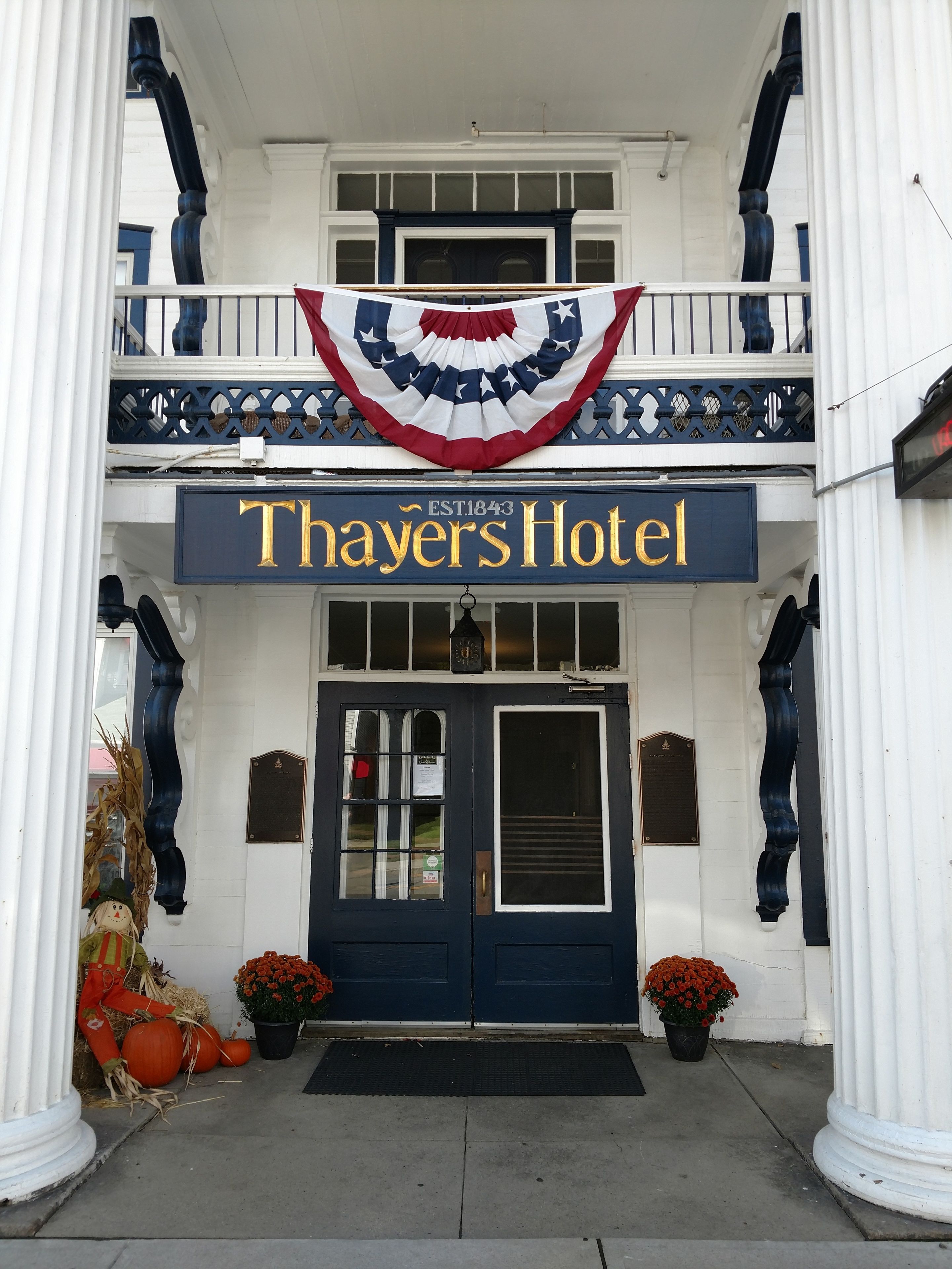 The front of Thayers Hotel with a prominent sign, a balcony with a decorative railing, and two potted plants with red flowers.