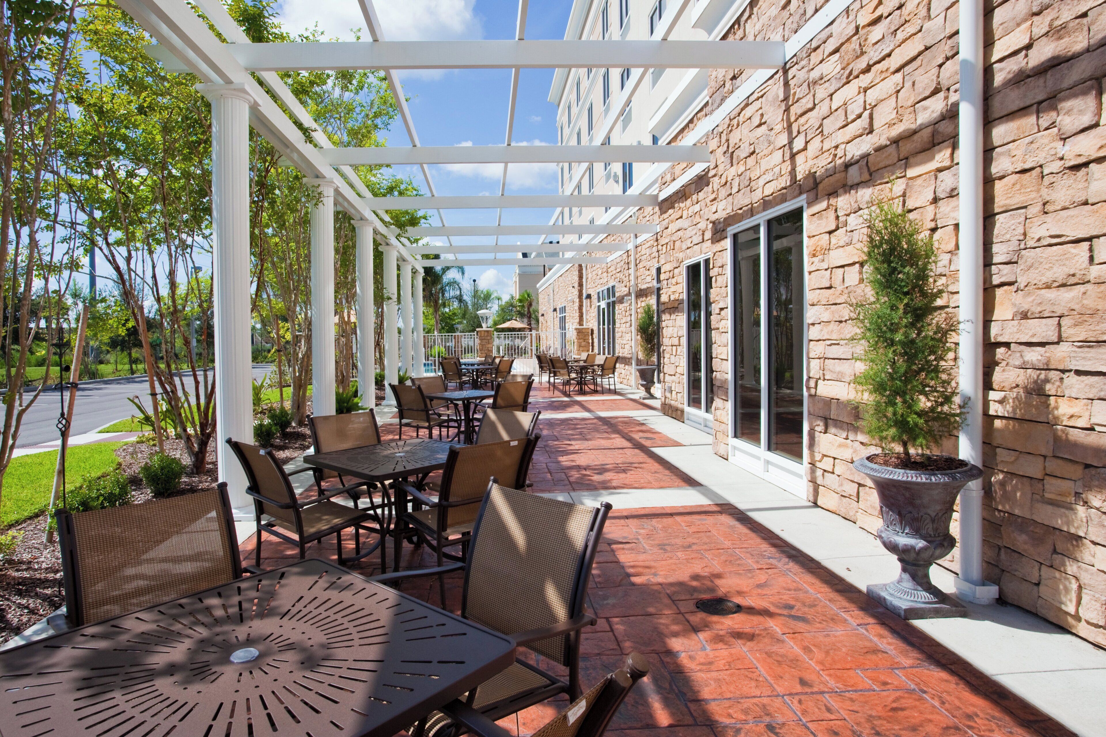 An outdoor patio with tables and chairs, a pergola, and a stone building with windows.