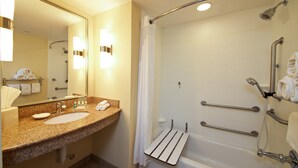 A hotel bathroom with a granite countertop sink, a large mirror, a shower with grab bars, and a towel rack.