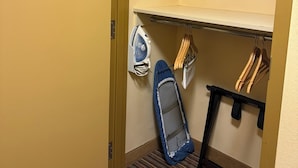 A hotel room with a striped carpet, a small ironing board, and a hanging rack with clothes.