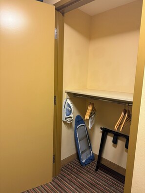Habitación de hotel con alfombra a rayas, una plancha pequeña y un perchero con ropa.