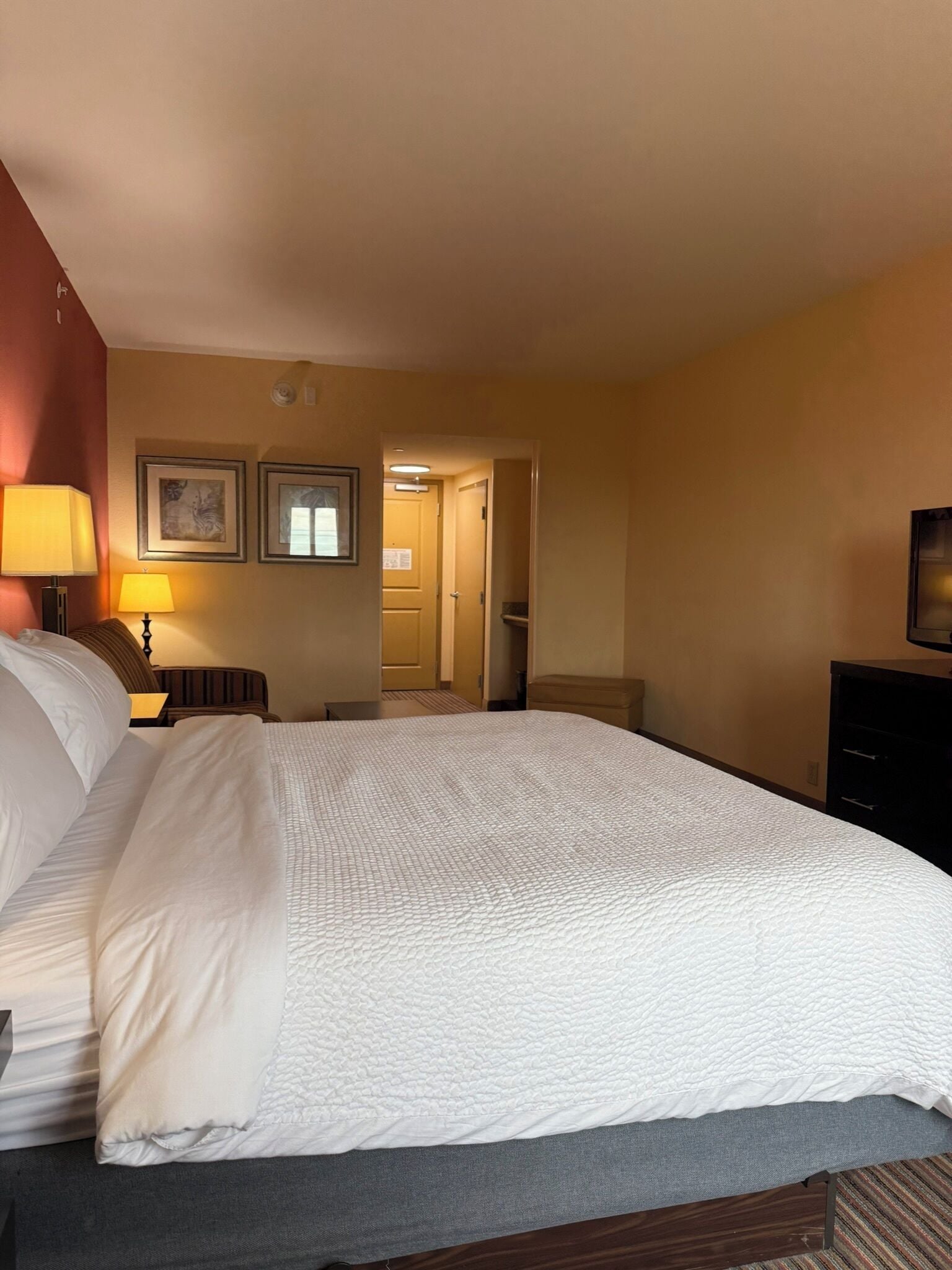 Habitación de hotel con una cama grande, una mesita de noche con lámparas, un aparador y una puerta que da a un pasillo.