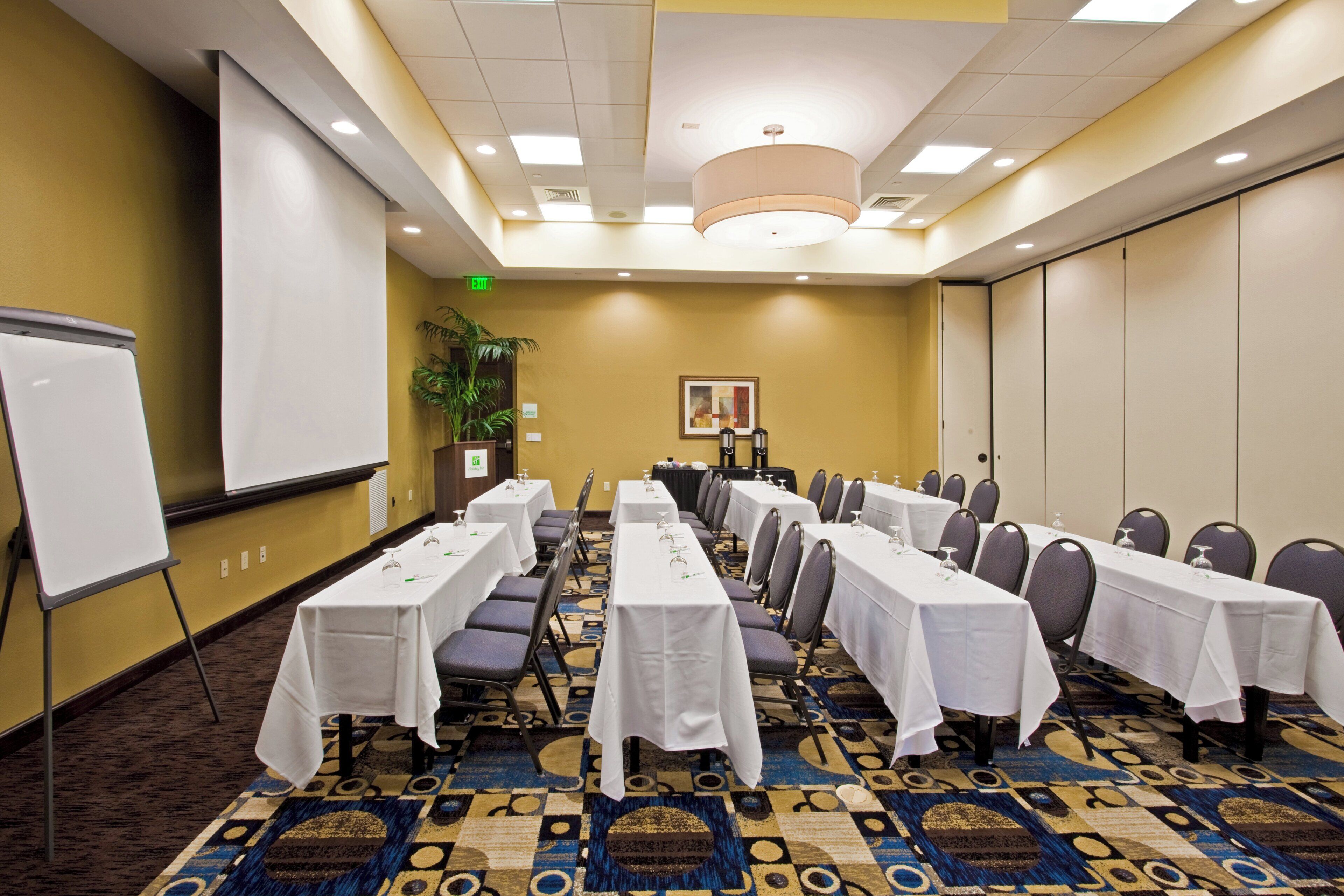 A conference room with long tables, chairs, a projector screen, and a carpeted floor.