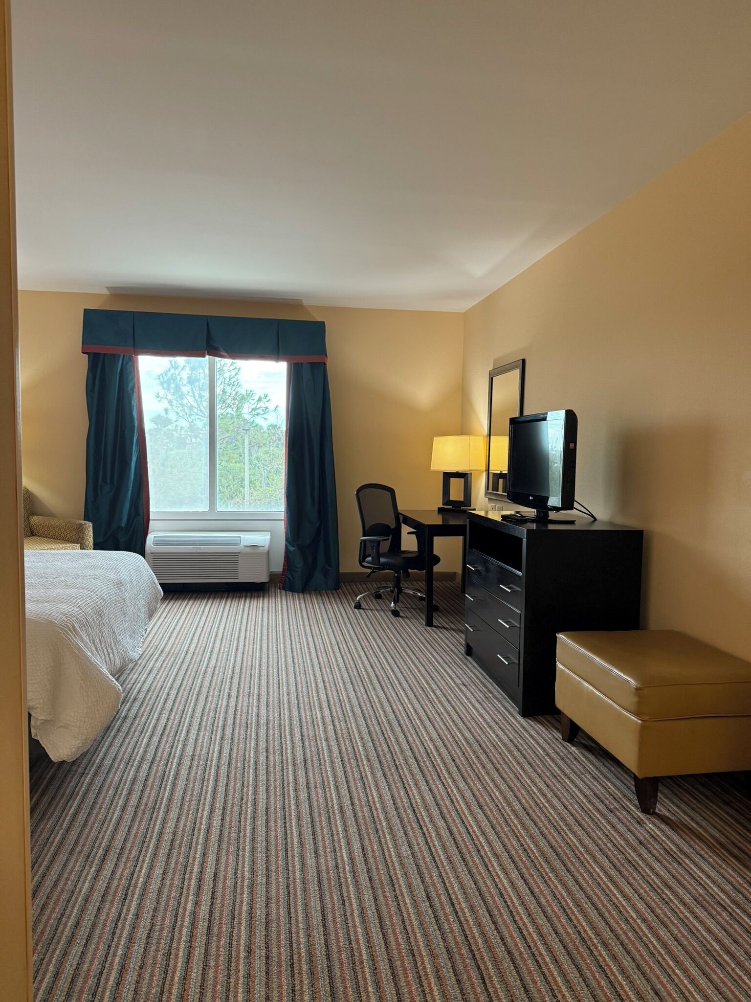 A hotel room with a bed, a desk with a computer, a chair, a dresser, and a window with curtains.